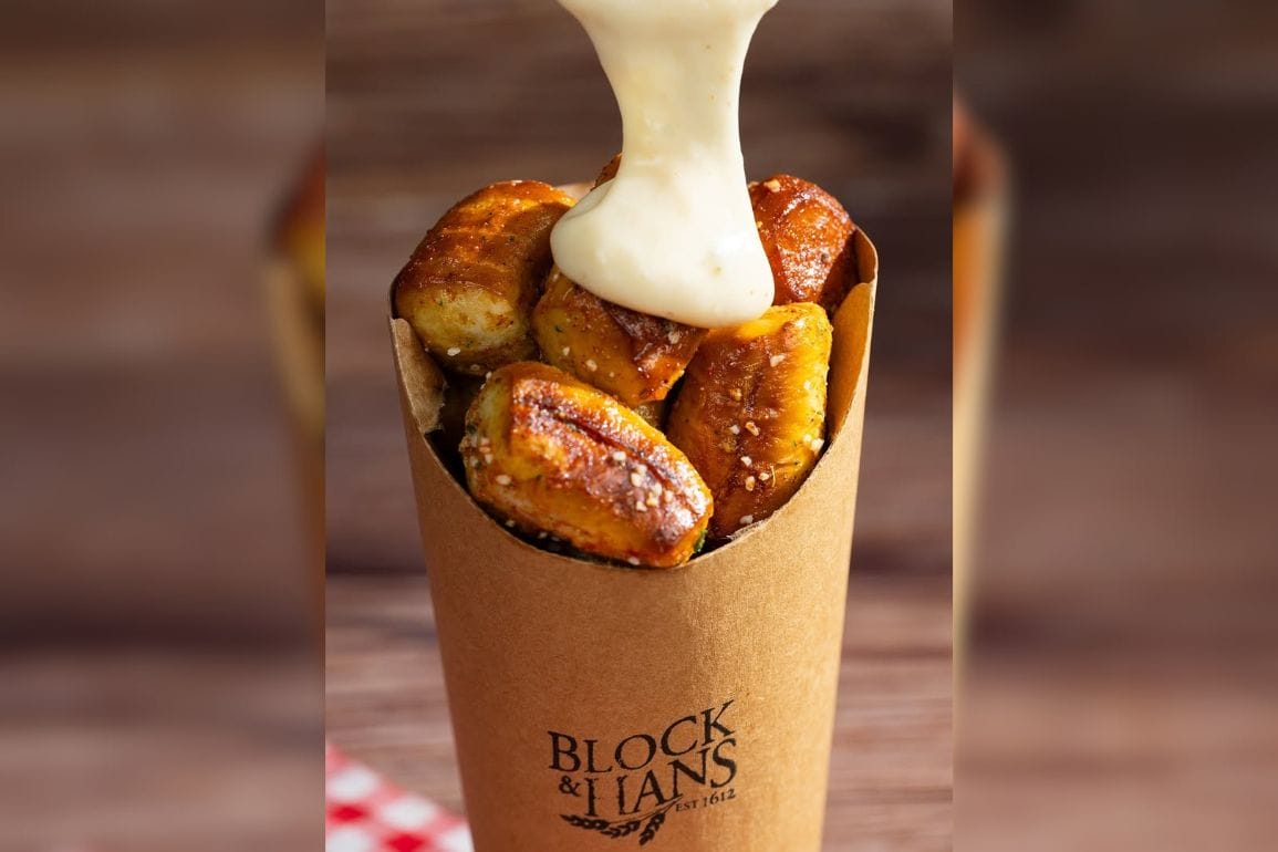 Delight in the irresistible charm of Block & Hans pretzel bites, nestled in a branded cardboard cup, as they're lovingly smothered with rich, golden cheese sauce cascading from above.