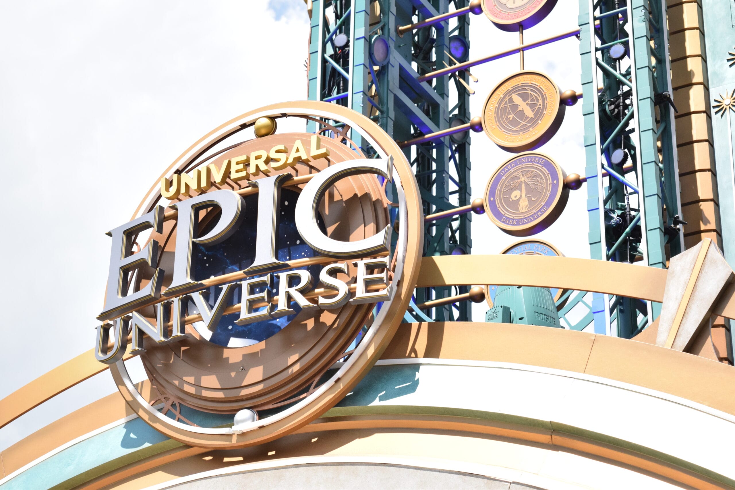 Universal Epic Universe logo on a theme park entrance structure, featuring circular emblems and decorative elements, invites guests into a world of wonder. As part of the Portals Tour, visitors will journey through new dimensions against a partly cloudy sky.