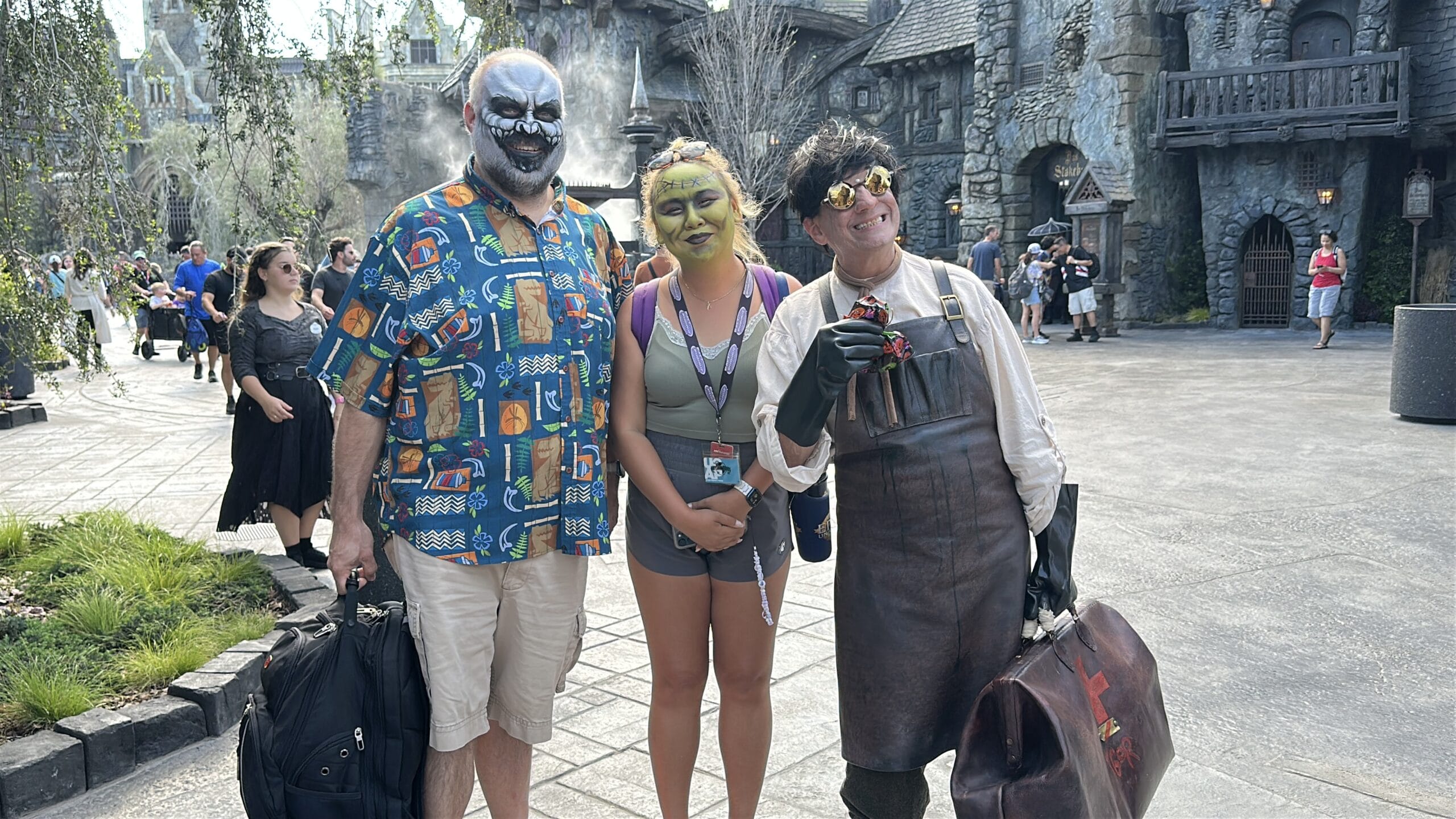 Three people in costume pose for a photo at an outdoor theme park; one shows off impressive Monster Makeup with clown flair, another is dressed as an ogre, and the third wears a lab coat with gloves and goggles. Perfect for a Darkmoor adventure!.