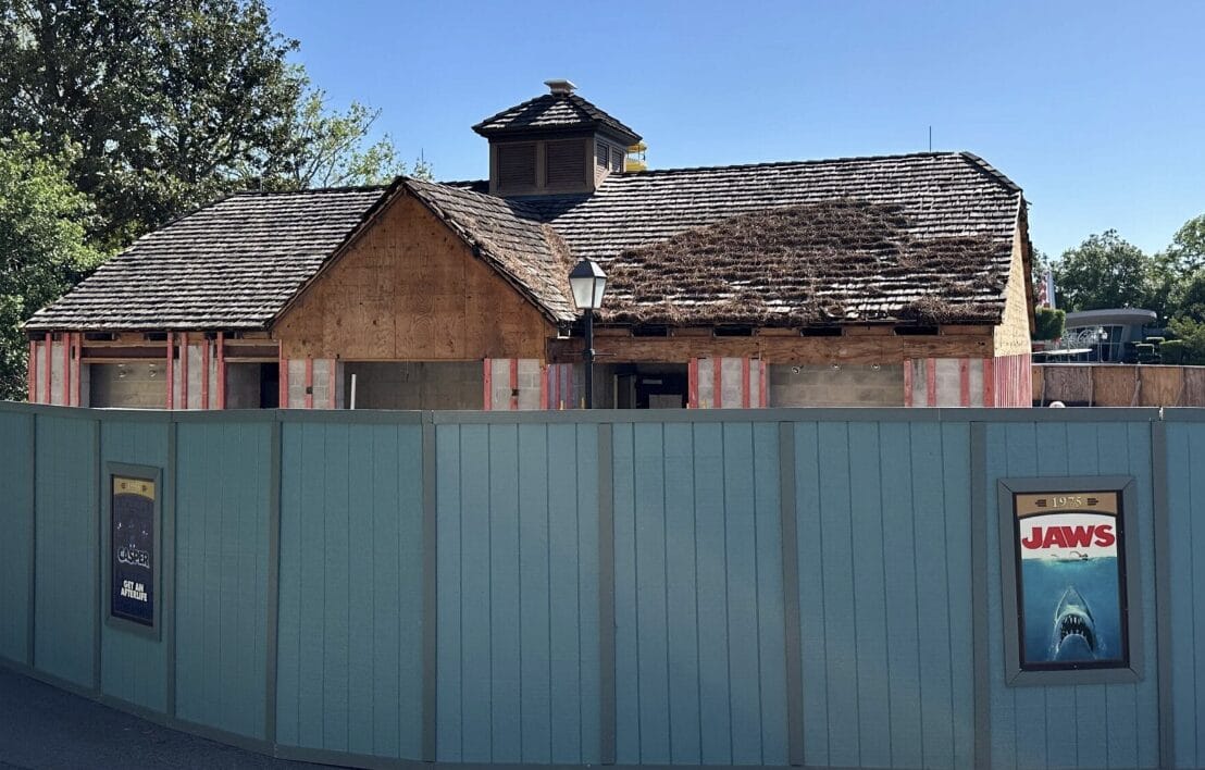 A wooden building under renovation, reminiscent of a Disney World Hotel, is partially obscured by a blue fence adorned with movie posters, including one for "Jaws." Trees and a clear sky enhance the backdrop.