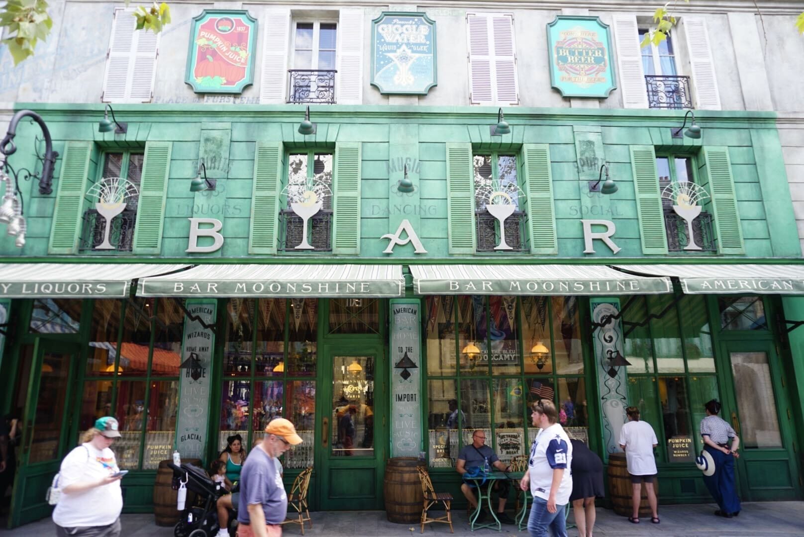People walk past Bar Moonshine, a green bar with large windows, signs, and decorative panels on a lively city street.