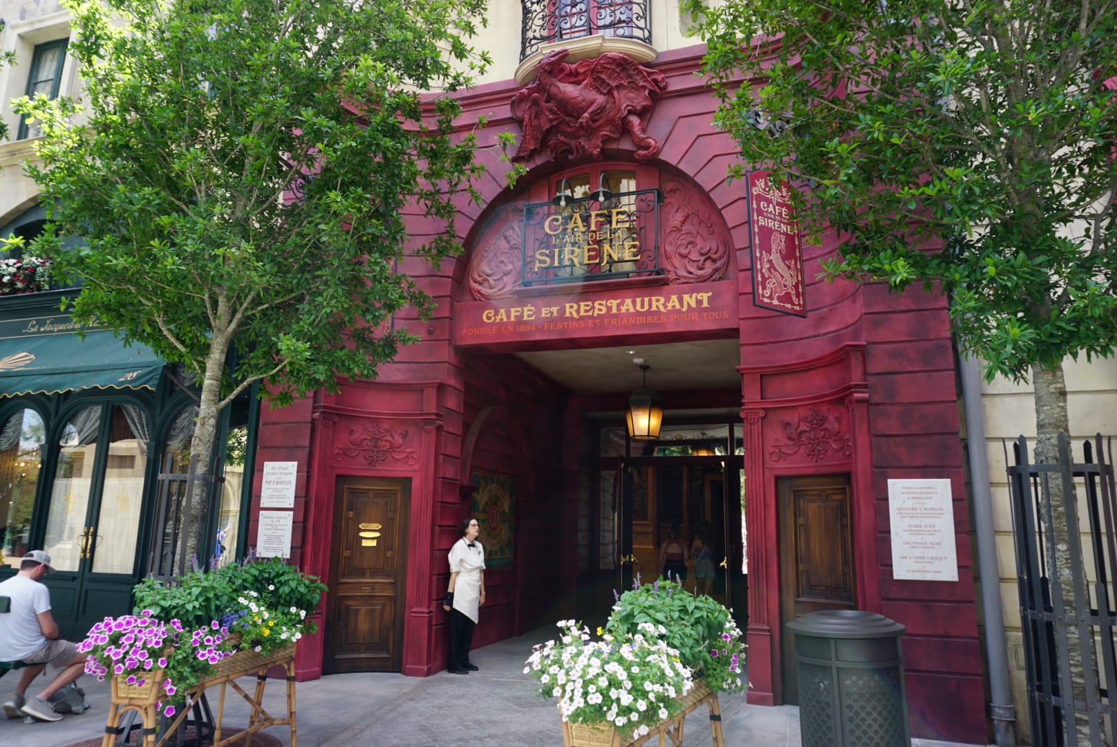Entrance to Café Sirène restaurant with red decor, flowers, and a staff member—see the full menu at the door.