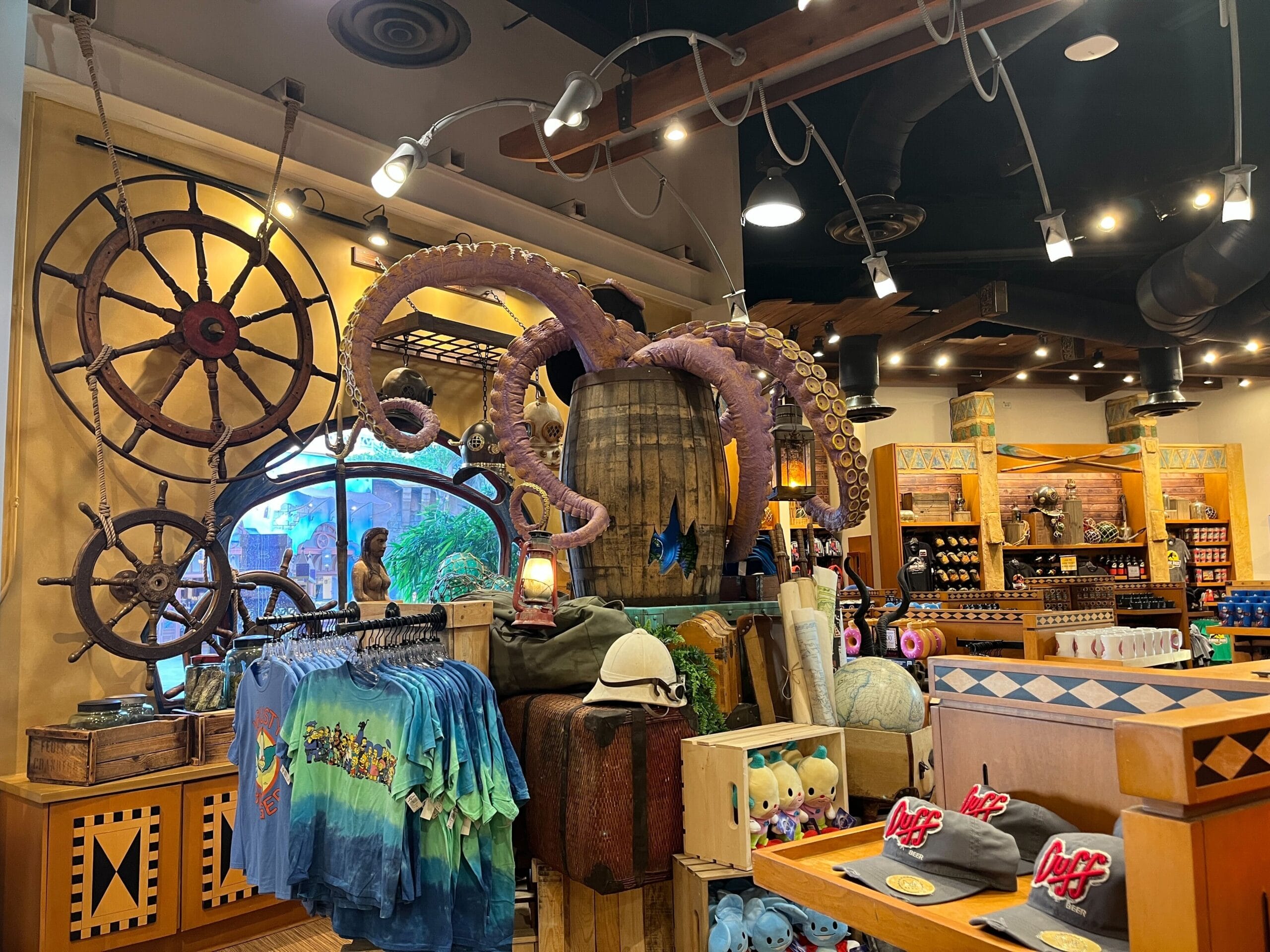 Inside the Trading Company Store at Universal Islands of Adventure, a nautical theme comes to life with new props—a large octopus sculpture wrapped around a barrel and ship's wheel. Display racks showcase shirts, hats, and an array of merchandise.