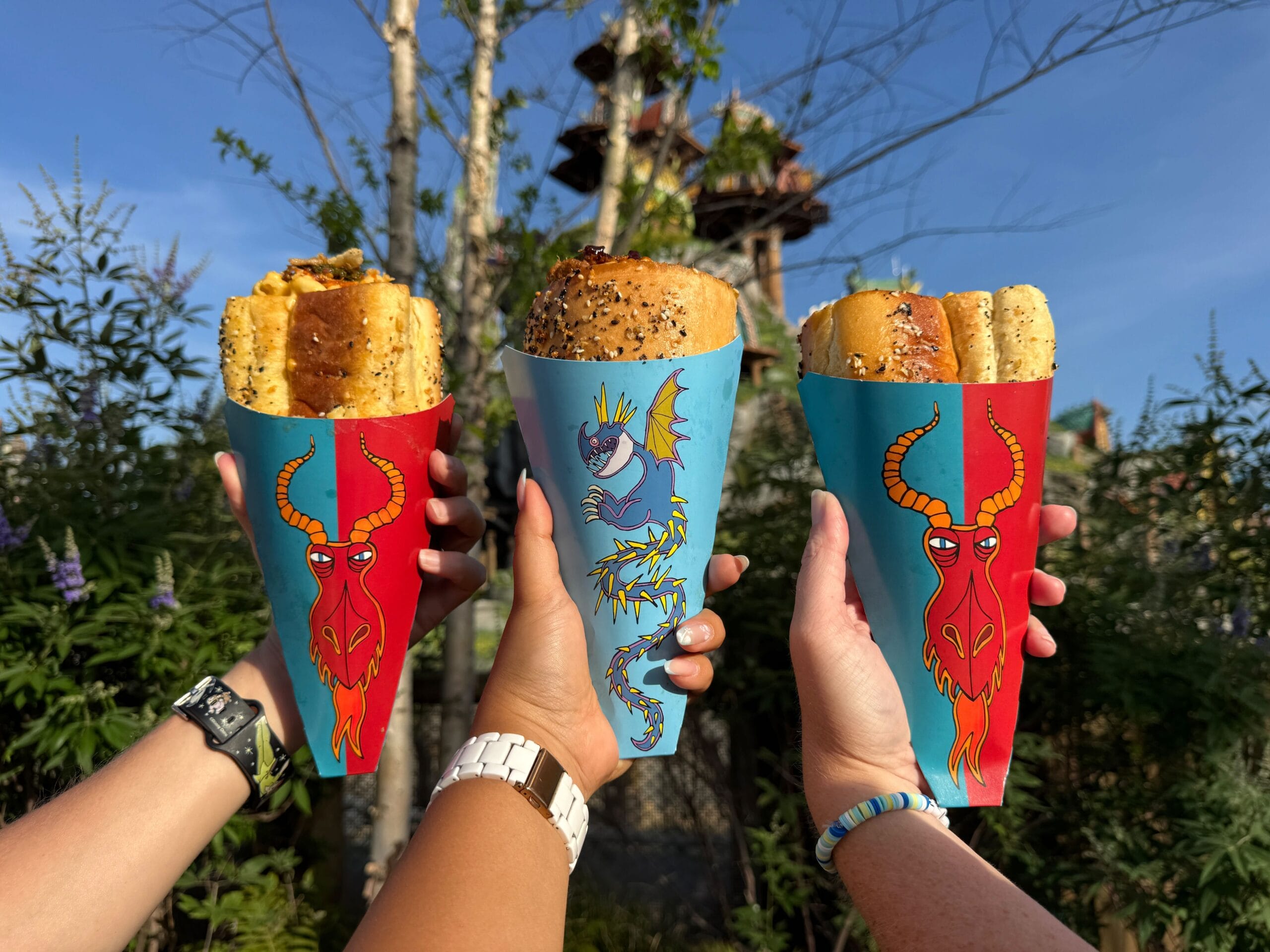 Three hands hold breadsticks in colorful cones from Hooligan's Grog & Gruel, with trees and a castle tower behind.