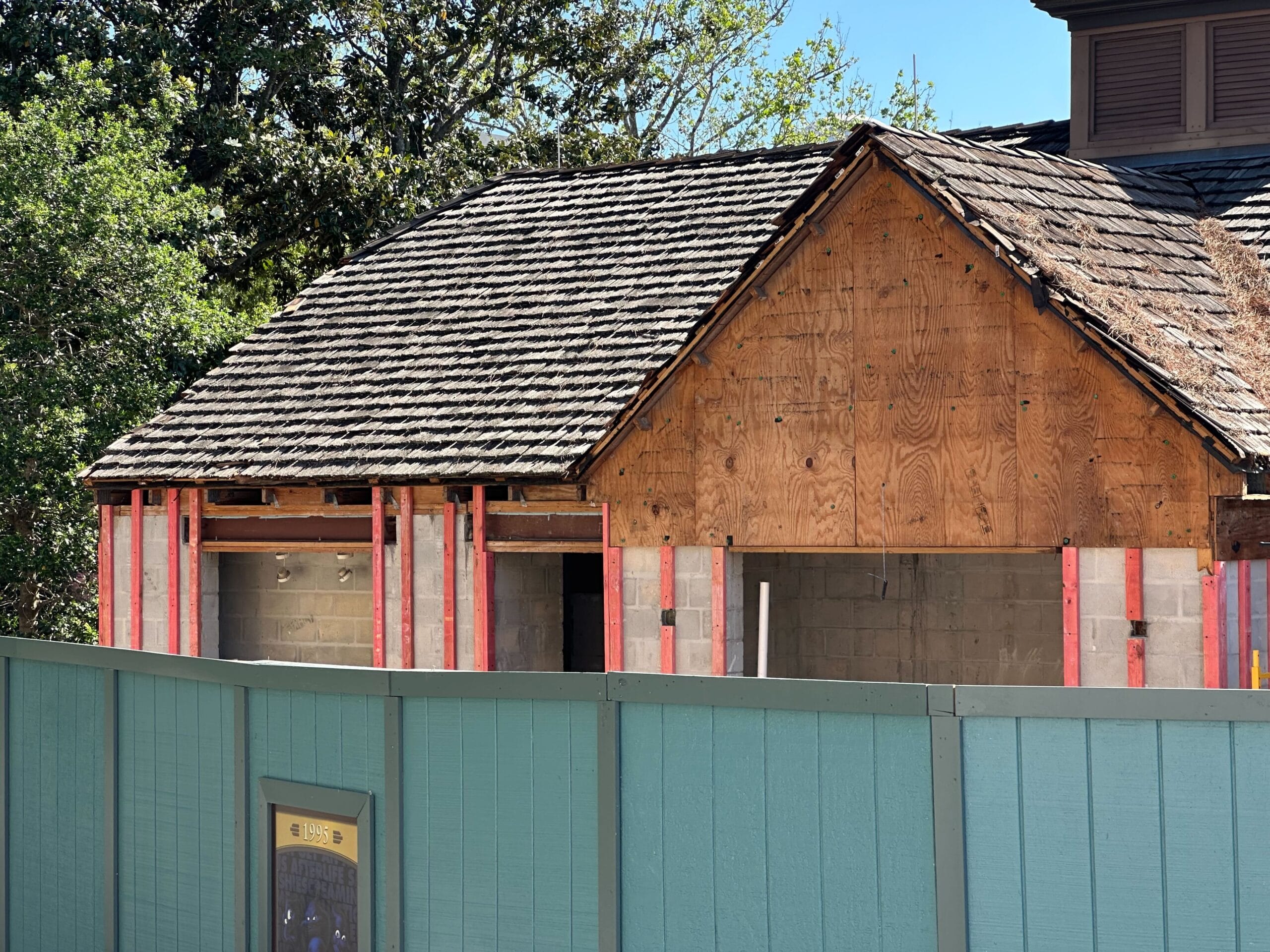 A small house under renovation with exposed wooden siding and a shingled roof. A green construction fence surrounds the site.