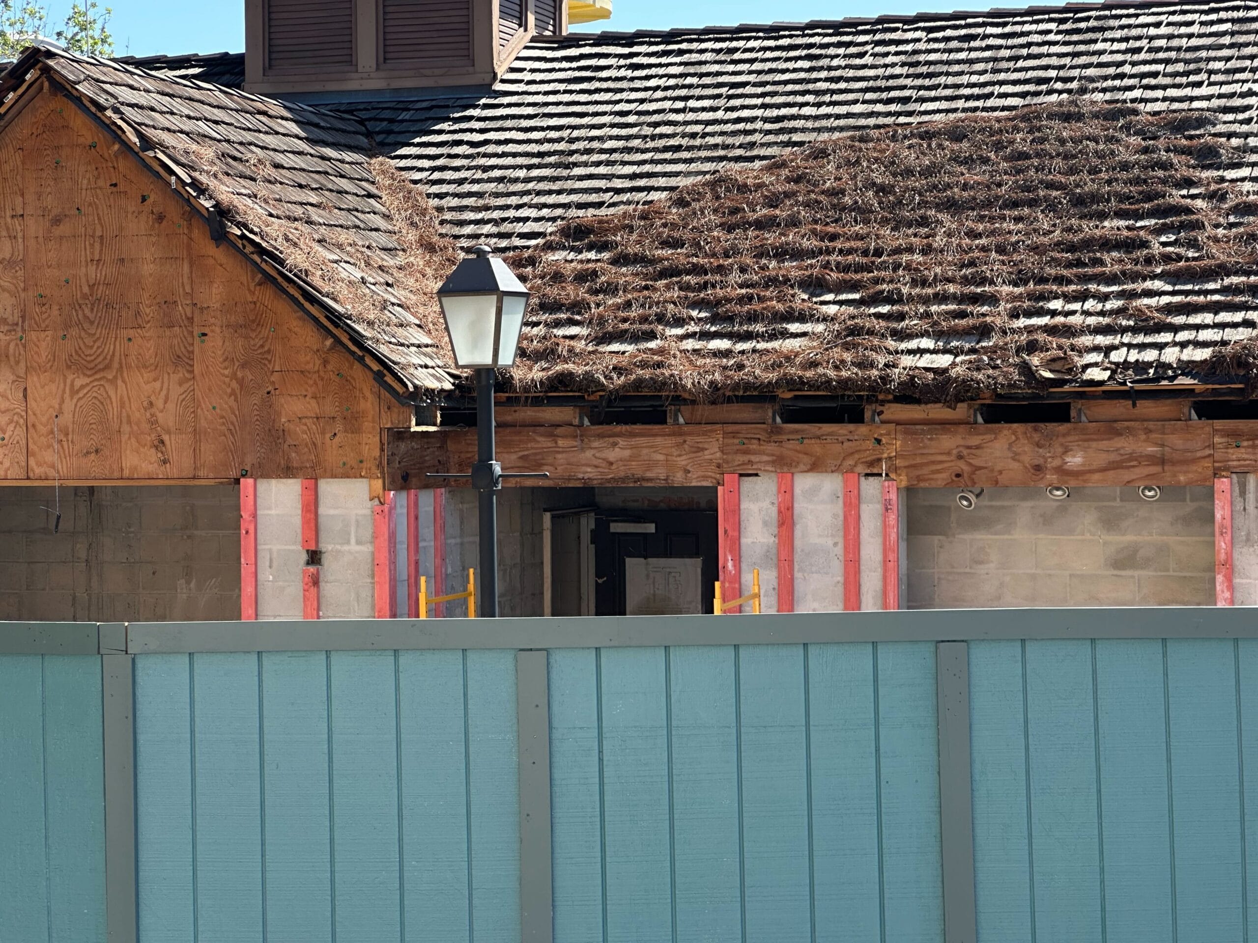 Partially demolished building with wooden roof and exposed wooden and cement walls. Light post in front, blue fence surrounds the site.