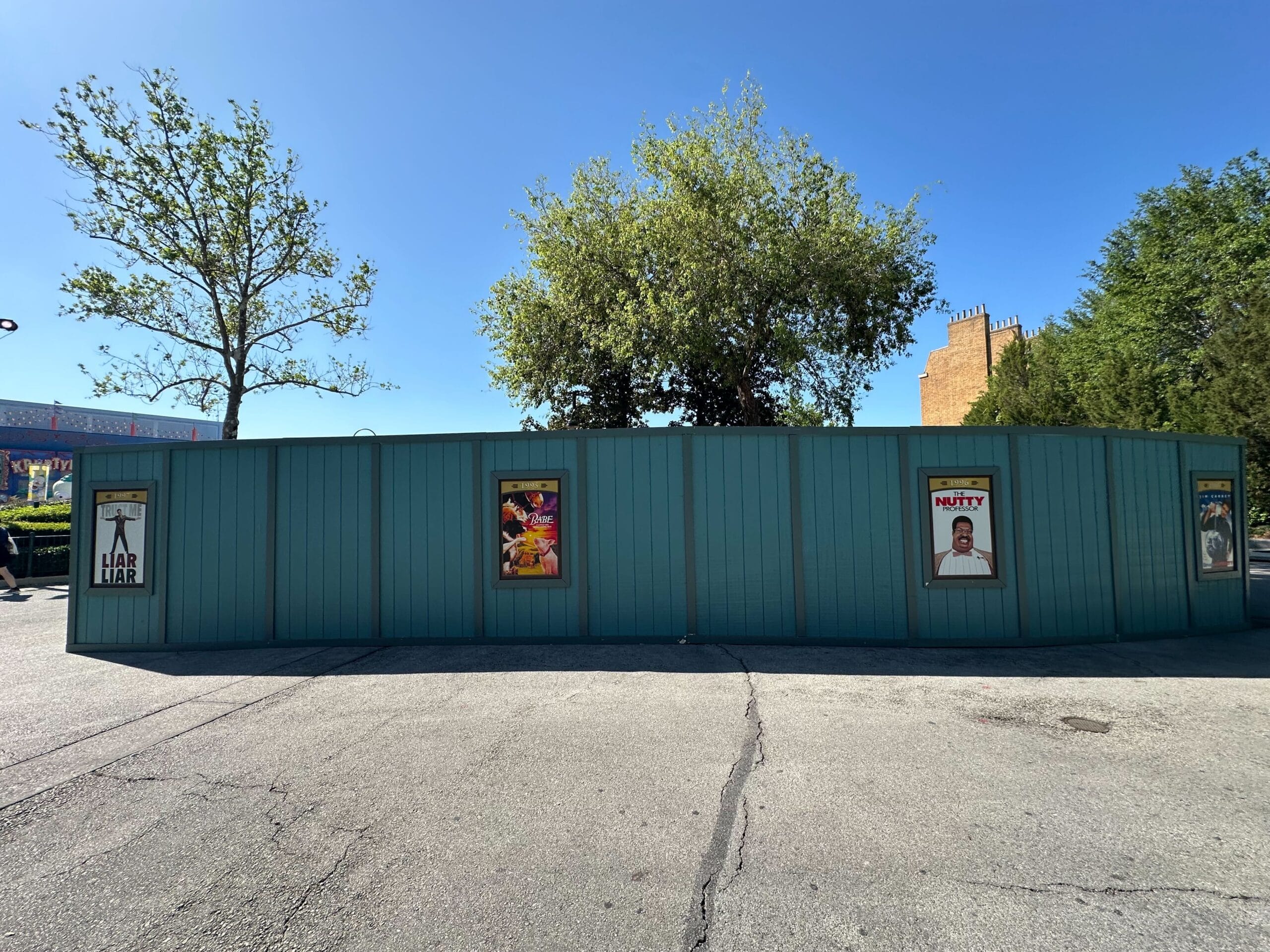 A teal wall with framed posters of various movies stands in front of trees and a clear sky. The ground is paved with light-colored asphalt.