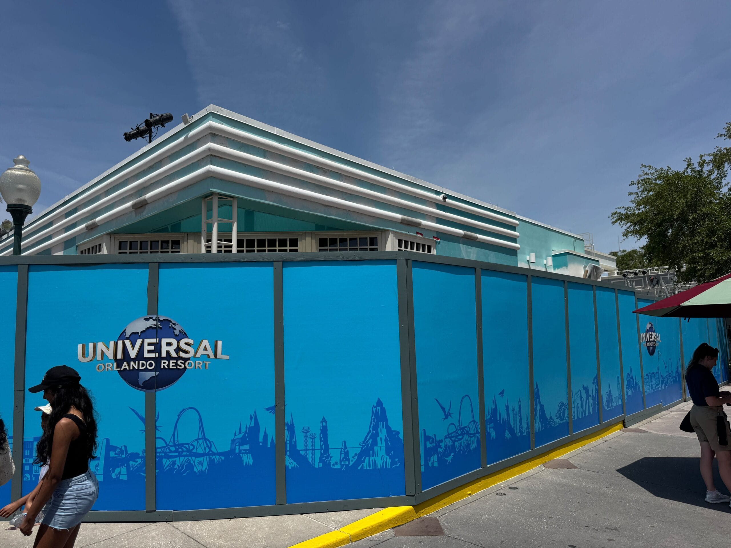 Blue construction walls signal shop construction at Universal Orlando Resort as two people walk by in the sunshine.