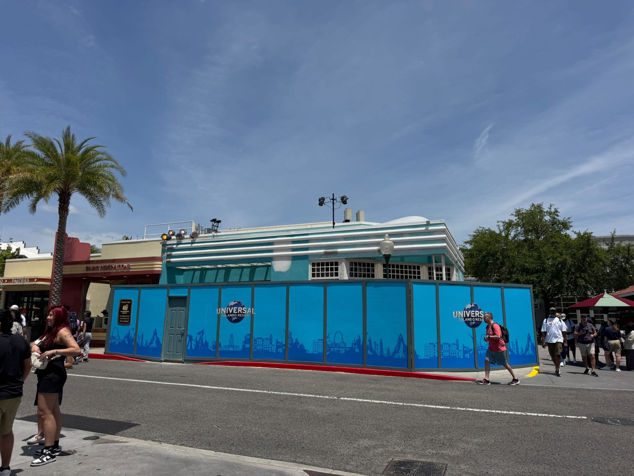 A blue shop construction wall with Universal Studios branding blocks a building as people walk nearby under a sunny sky.