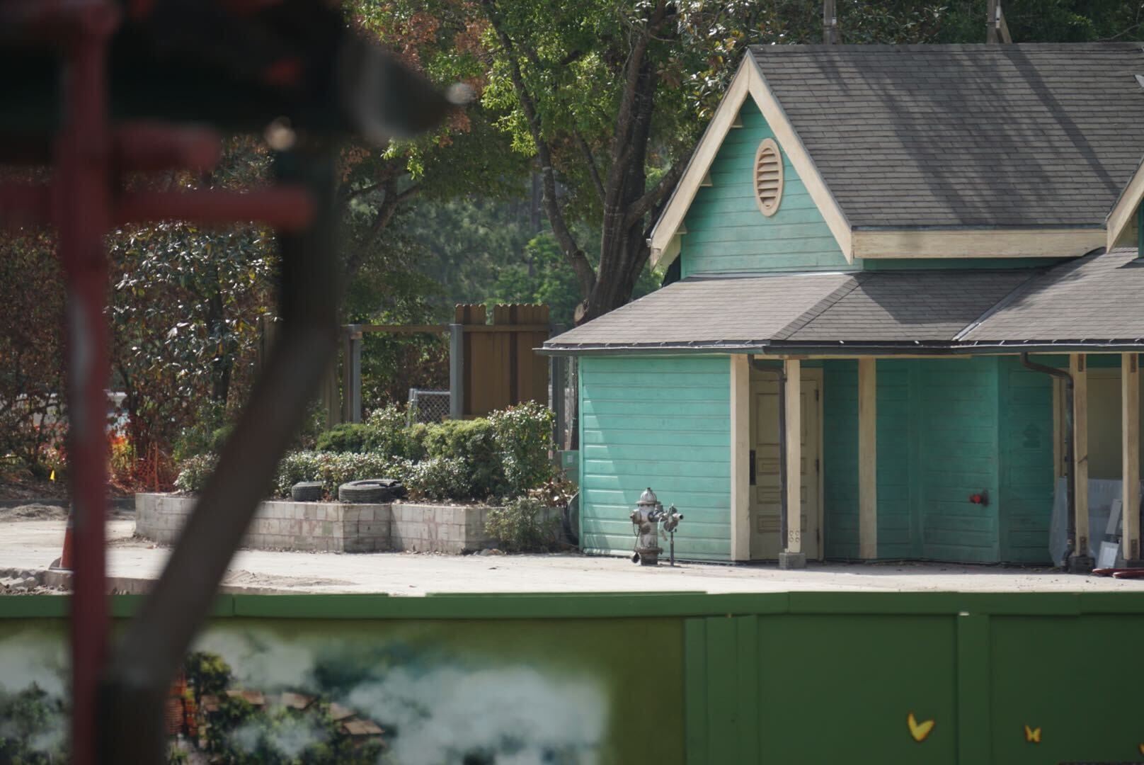 A green house with a sloped roof, tropical trees behind, and a fire hydrant by the path visible out front.