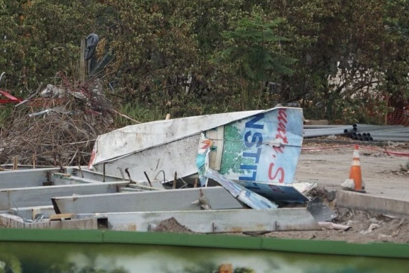 The construction site at DinoLand U.S.A. features debris, metal framework, and caution tape. Trees stand silhouetted in the background while a partial billboard peeks through, hinting at the prehistoric-themed adventure awaiting beyond the chaos.