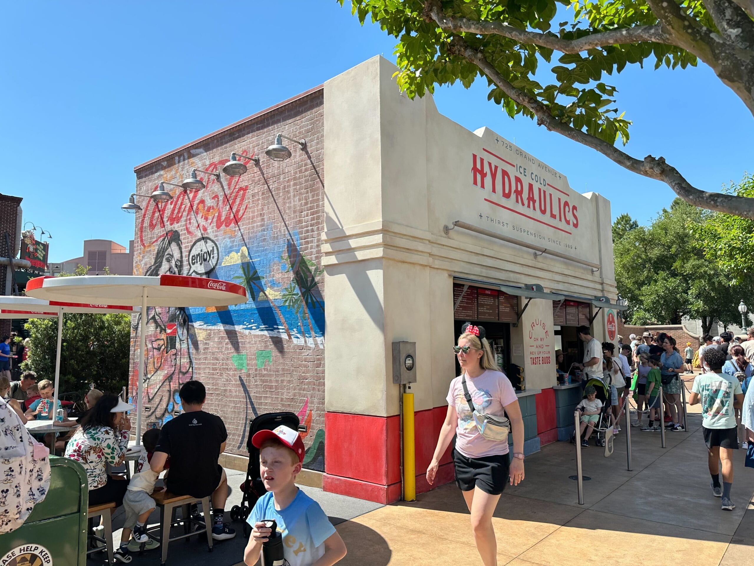 People sit and walk near the Ice Cold Hydraulics food stand with a Graffiti Style Mural on a sunny day.