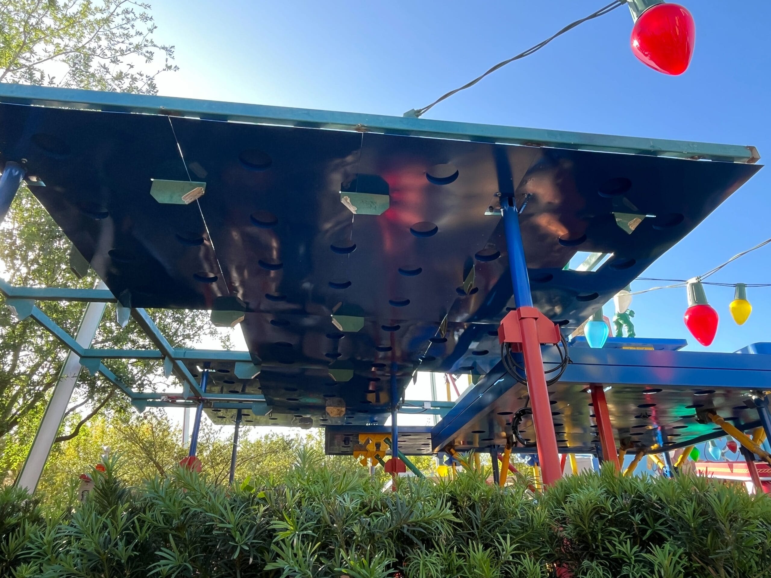 Underside view of a large playground structure resembling a board game piece, surrounded by greenery. Strings of oversized multicolored Christmas lights are visible above.