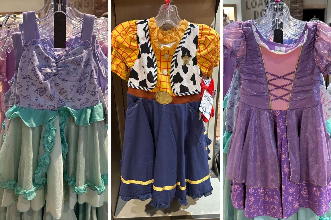 On hangers are three enchanting story play dresses: a green and purple dress adorned with unicorns, a cowboy-themed dress featuring a cow print vest, and a whimsical purple and pink dress with a lace-up front.