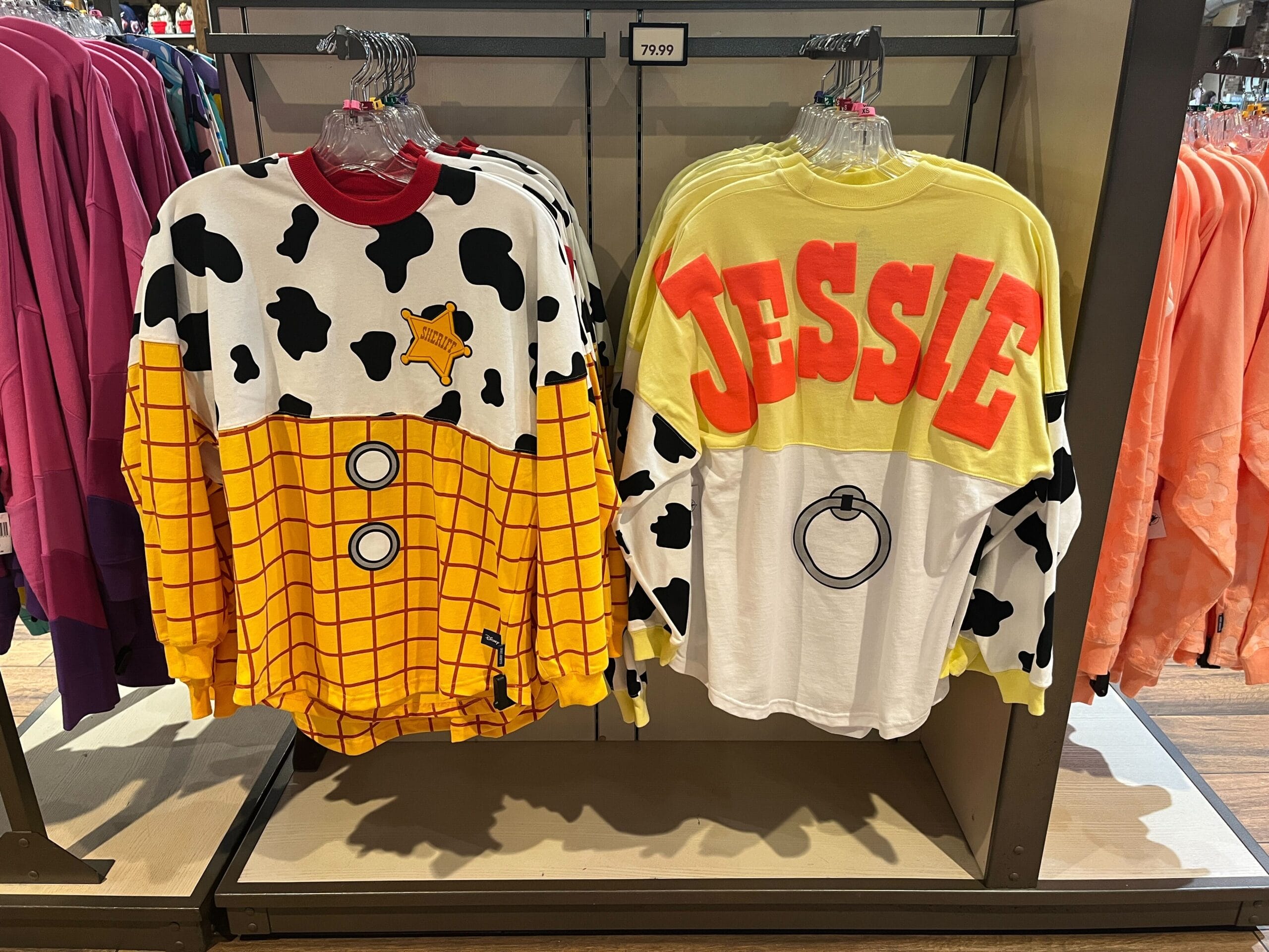 Two themed sweatshirts hang on a rack. One resembles a sheriff's outfit with cow print and badge; the other features large "JESSIE" text with red accents and a cow print.