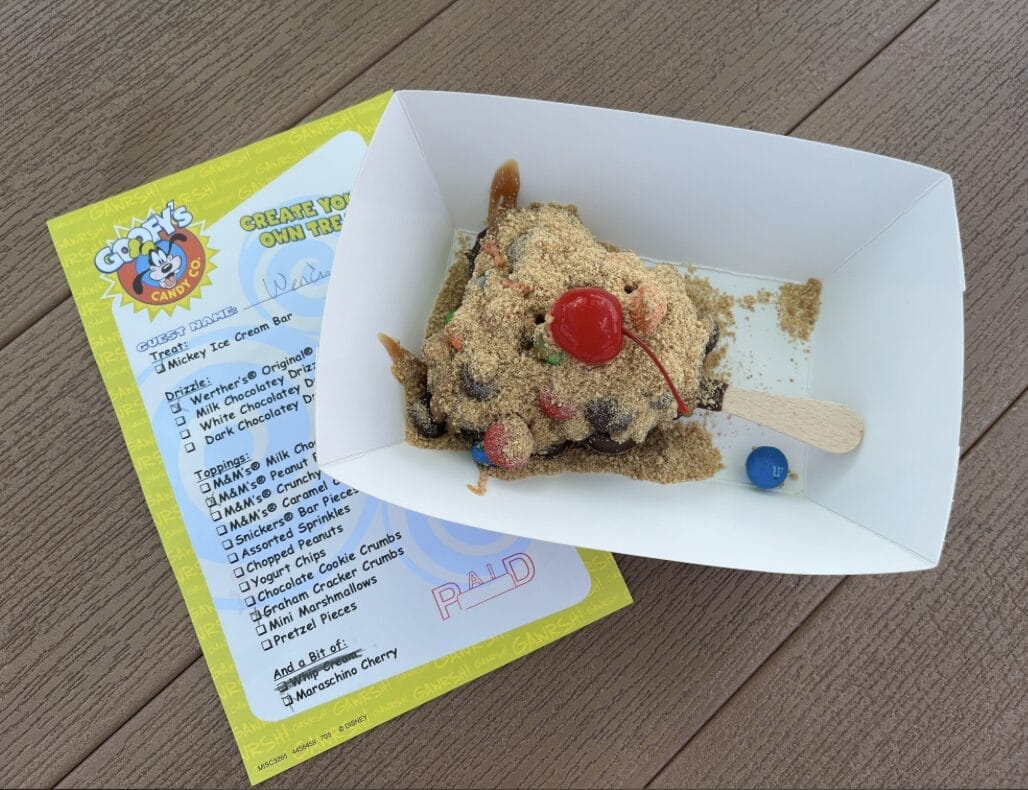 Goofy's Candy Co. presents an ice cream bar with a rich brown topping and a cherry crown, nestled in a paper tray beside a vibrant yellow create-your-own-treat checklist with marked options, resting atop a wooden surface adorned with a playful blue bead.