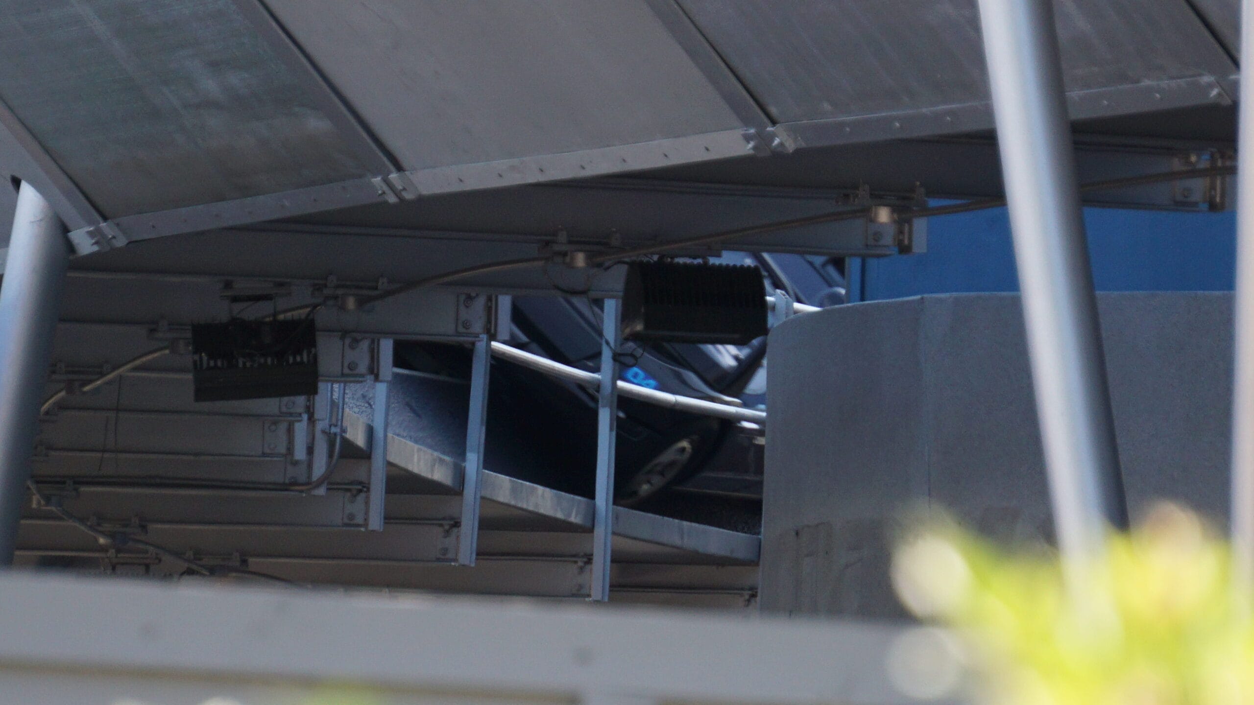 A car is partially visible under a metal structure, with its front peeking out near a concrete pillar and cables overhead.