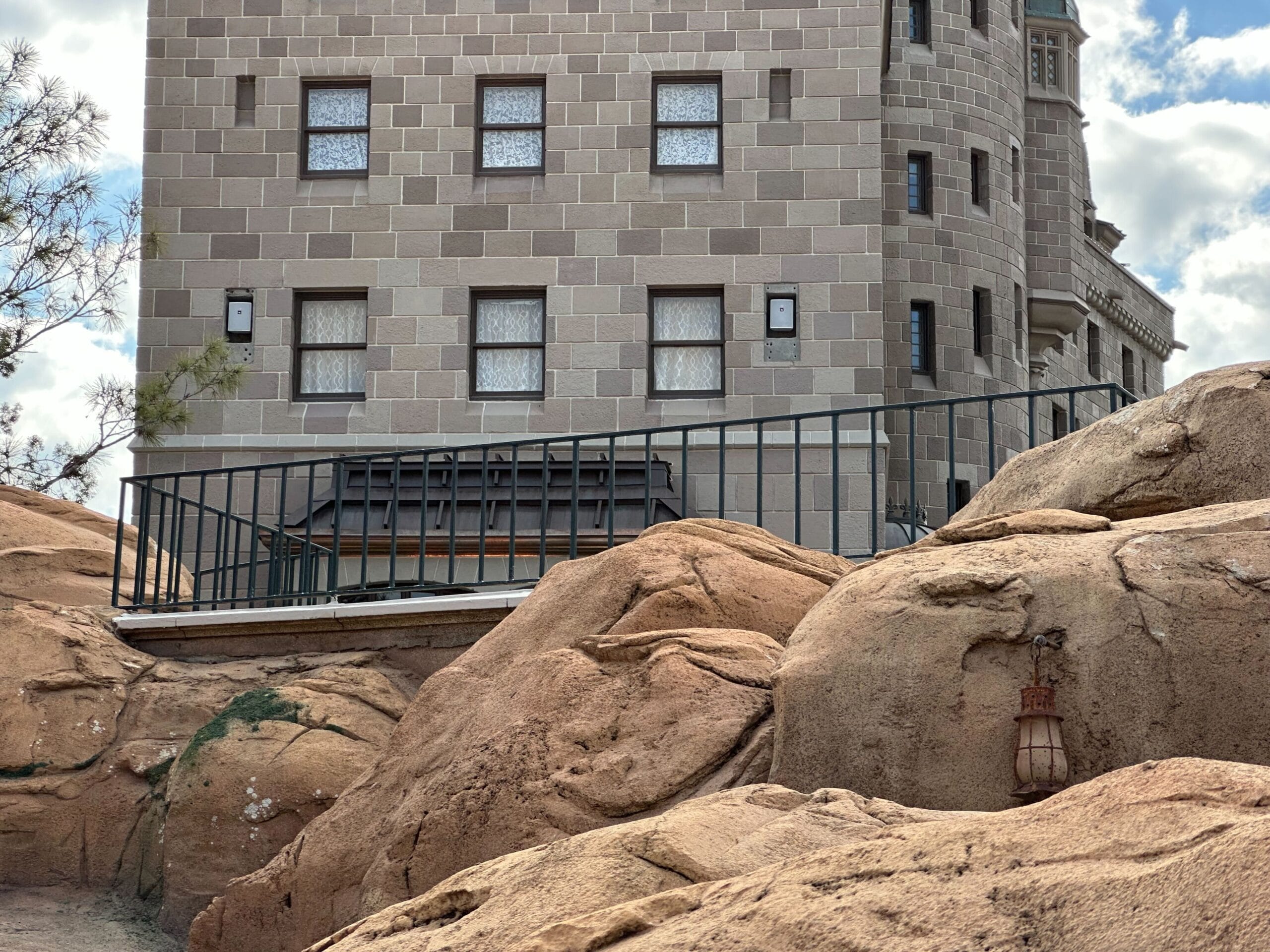 A stone building with small windows and balcony sits behind a metal railing and large tan boulders; a light fixture is attached to one rock.