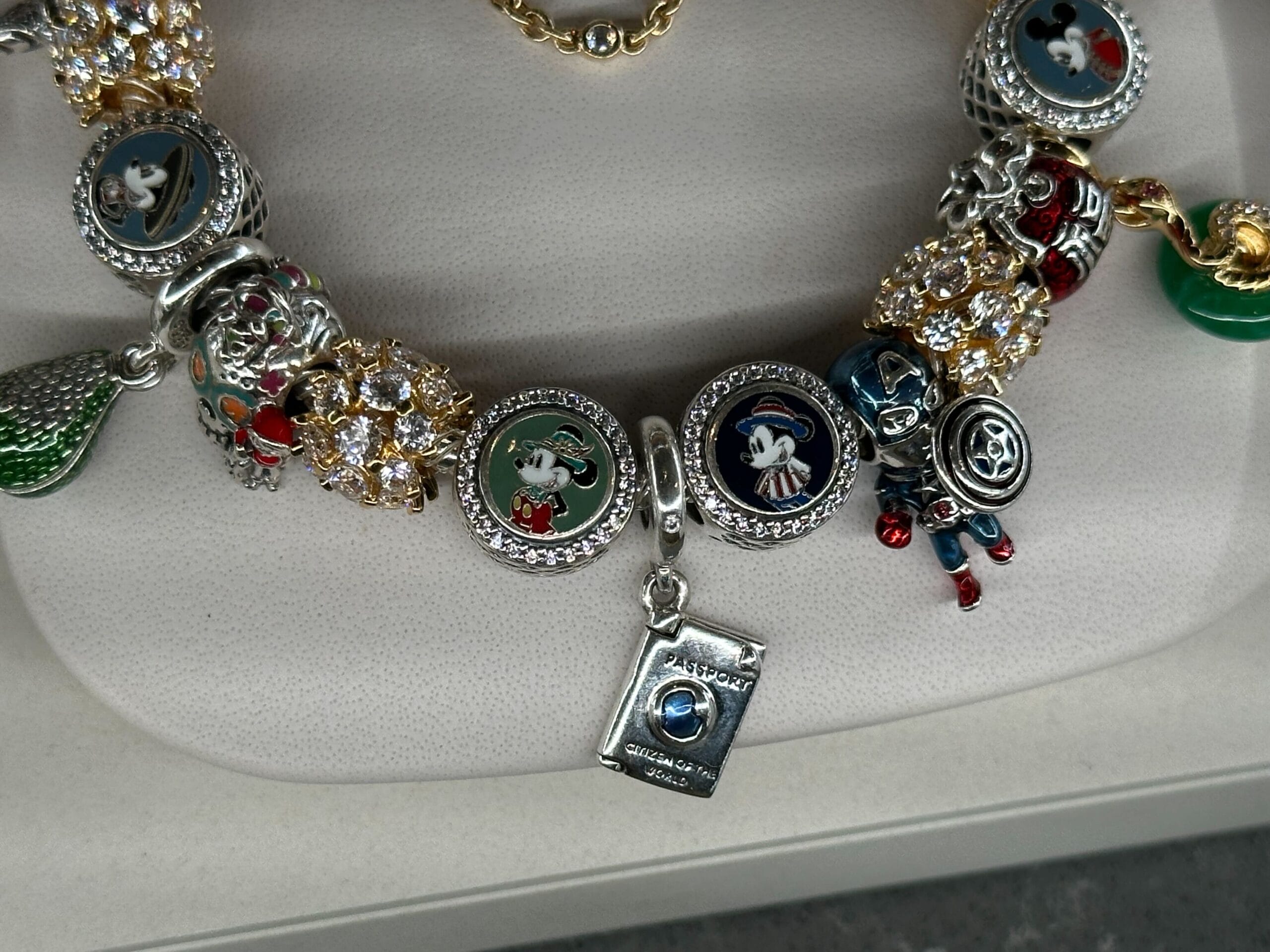 A bracelet adorned with various metal charms, including cartoon characters, a passport, and decorative beads, displayed on a white cushion.