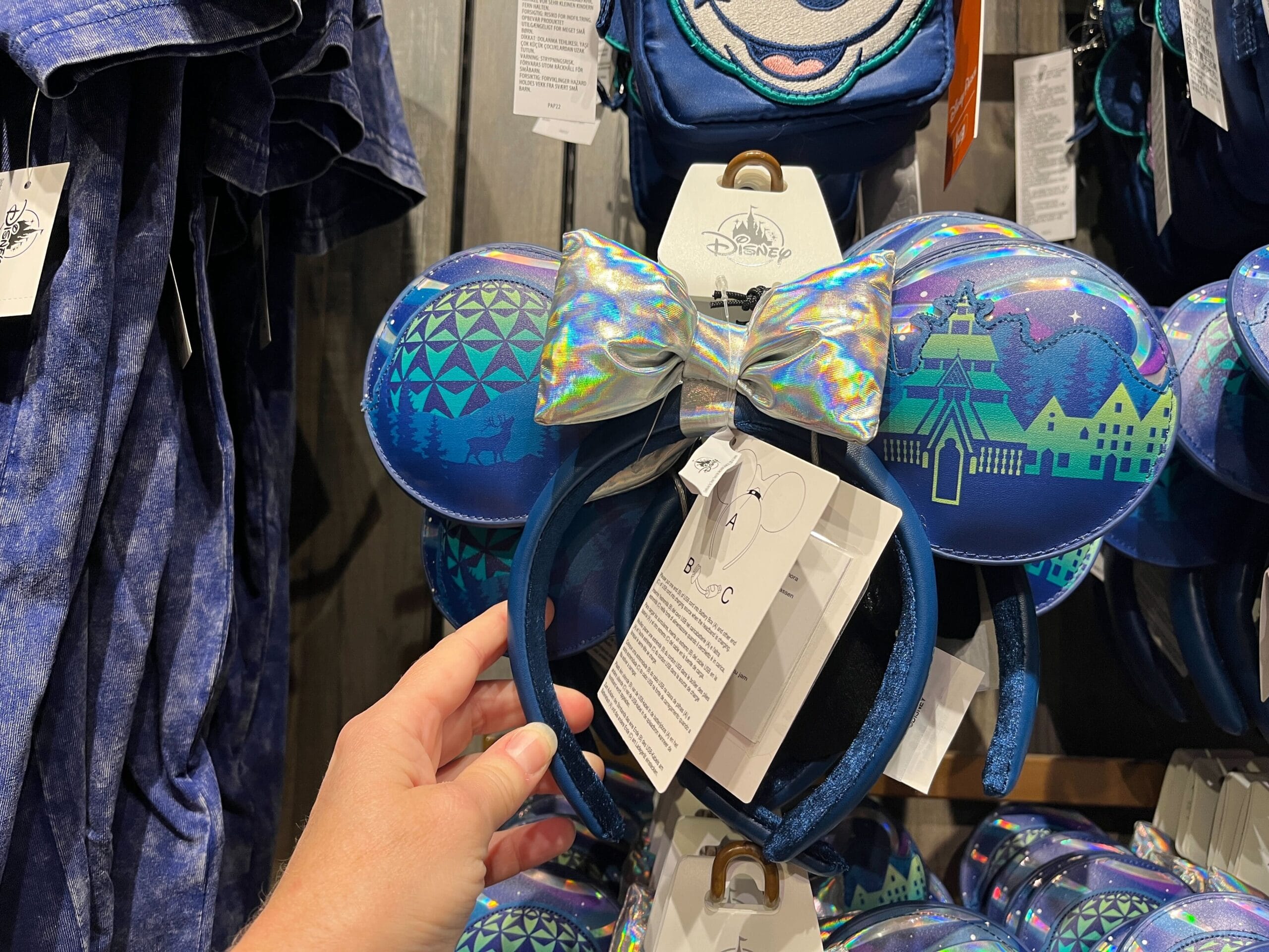 A hand holds an EPCOT-themed headband with blue ears showcasing geometric and landscape designs, complemented by a shiny silver bow. This Pavilion-inspired accessory is displayed alongside other clothing items in a store.