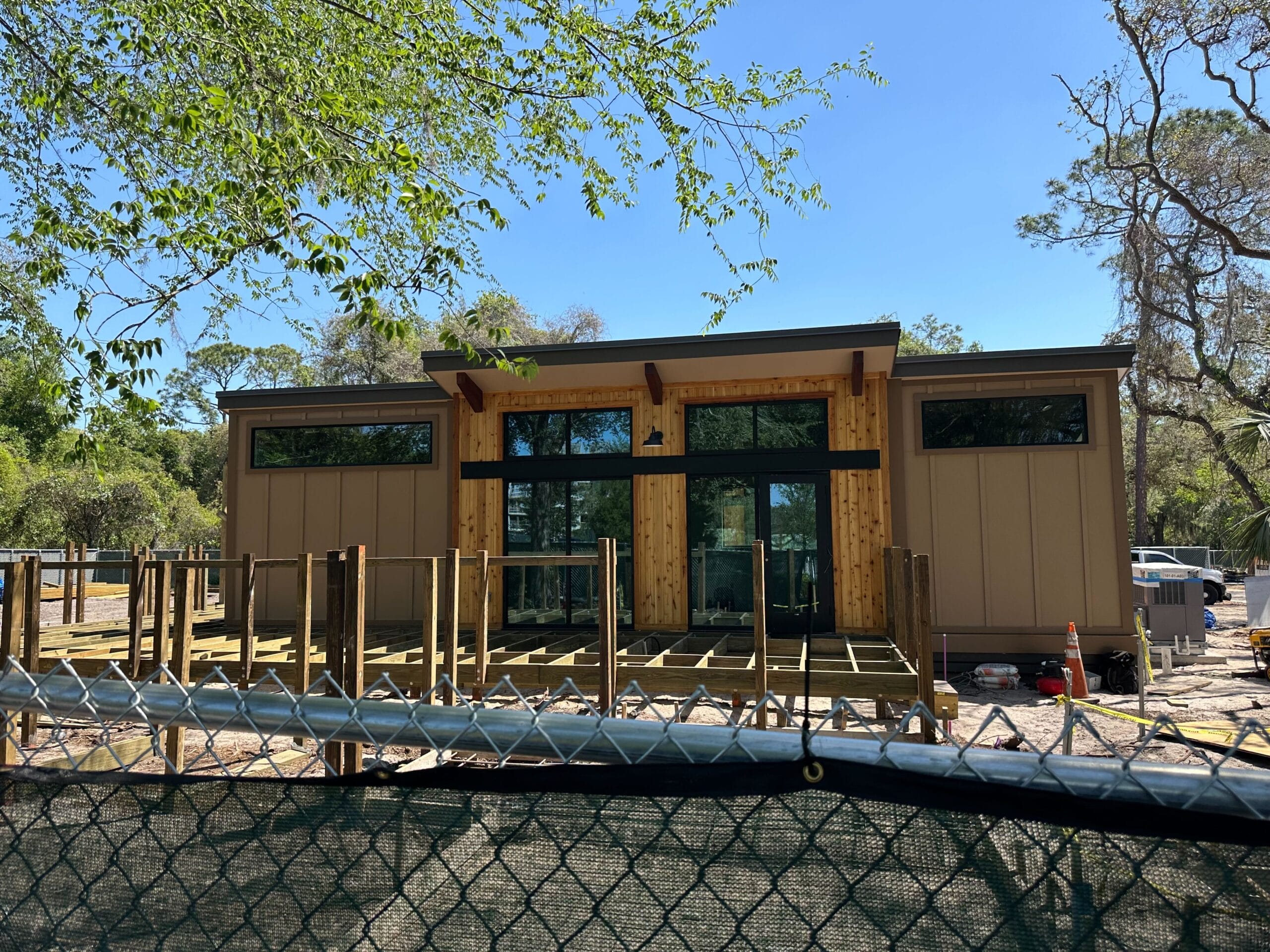 A small modern building under construction, featuring large windows and wooden accents reminiscent of Disney's Fort Wilderness Resort. Decks framed by the structure hint at future outdoor spaces. A chain-link fence surrounds the site, with trees in the background under a clear blue sky.
