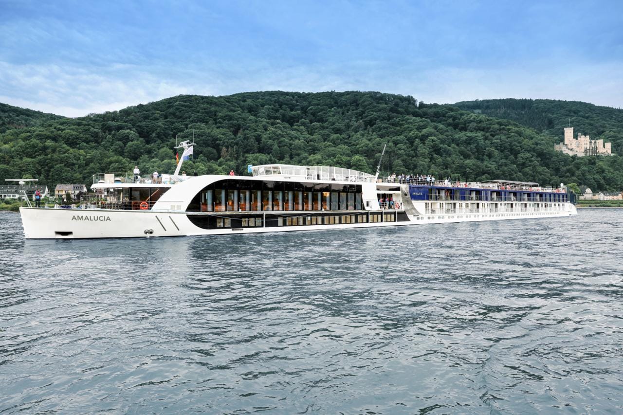 A large white cruise ship, offering exclusive perks like complimentary pre-night stays for Disney Visa Cardmembers, sails gracefully on a river with forested hills and a majestic castle in the background under a clear blue sky.