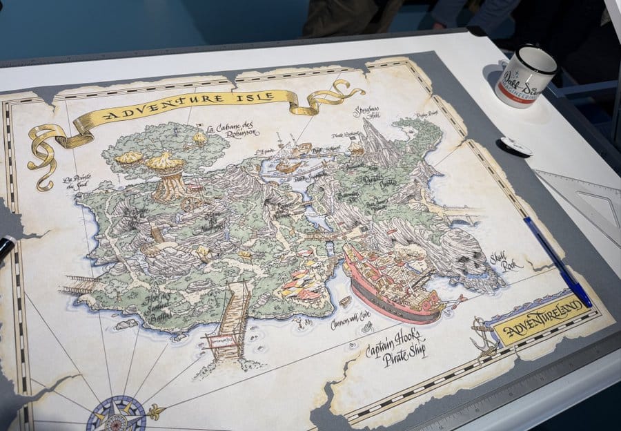 Illustrated map of Adventure Isle on a table, featuring landmarks like Captain Hook's Pirate Ship, and surrounded by art supplies including a pen and a mug.