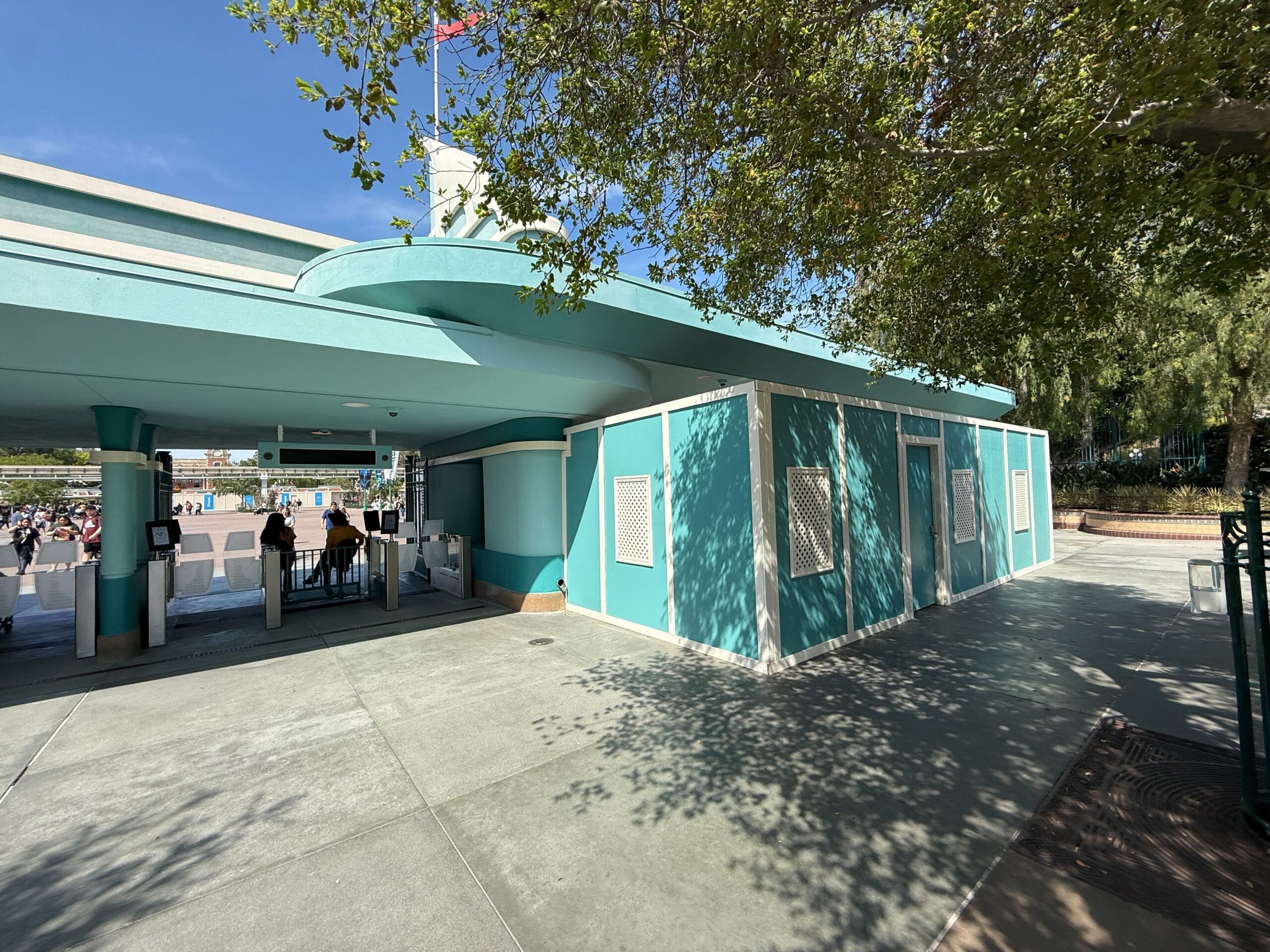 Entrance to a theme park with turquoise structures, turnstiles, and shaded area from nearby trees. A few people are seen in the distance.