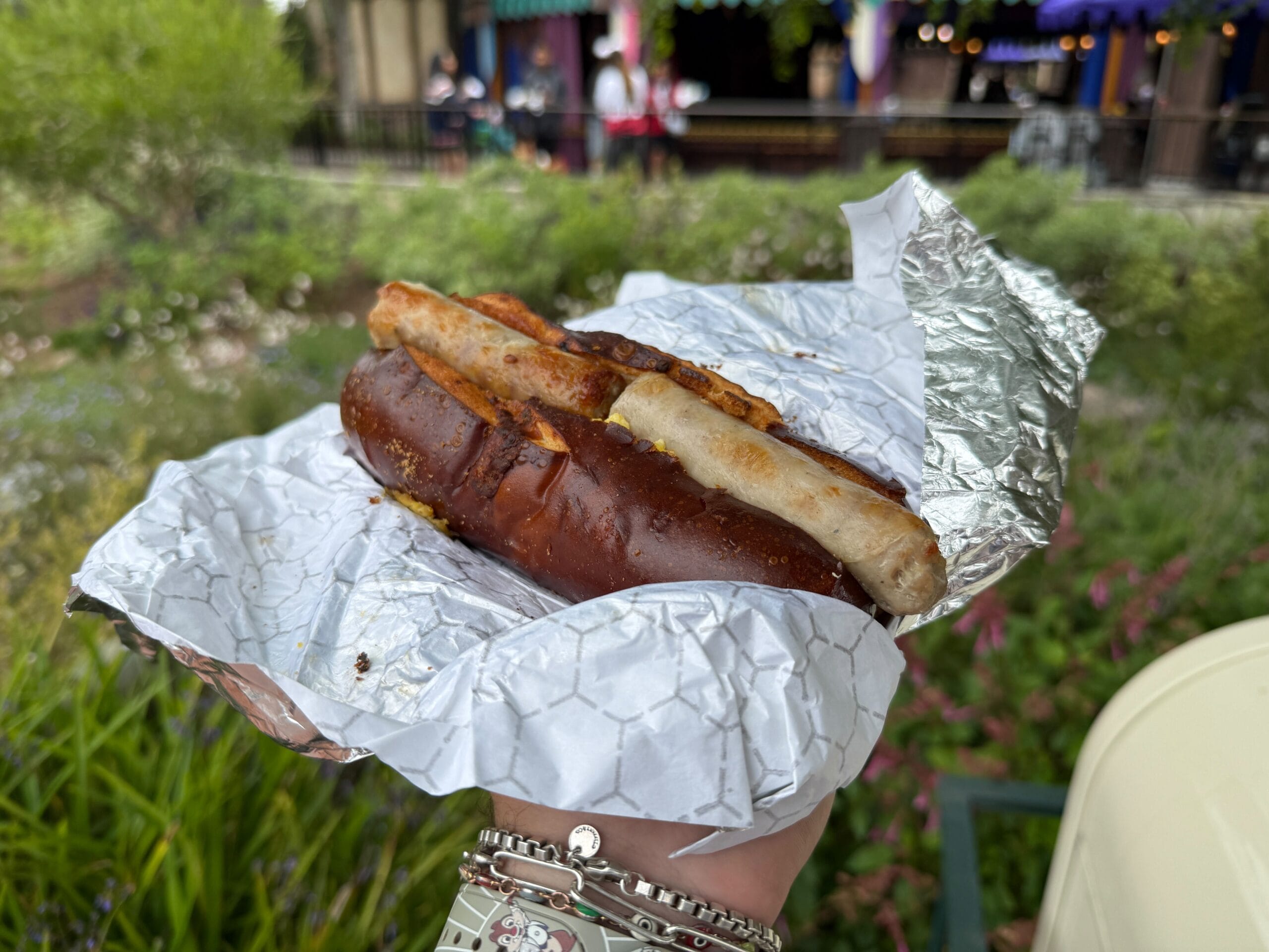 A hand holds a pretzel bun sausage wrapped in foil, with greenery and buildings blurred in the background.
