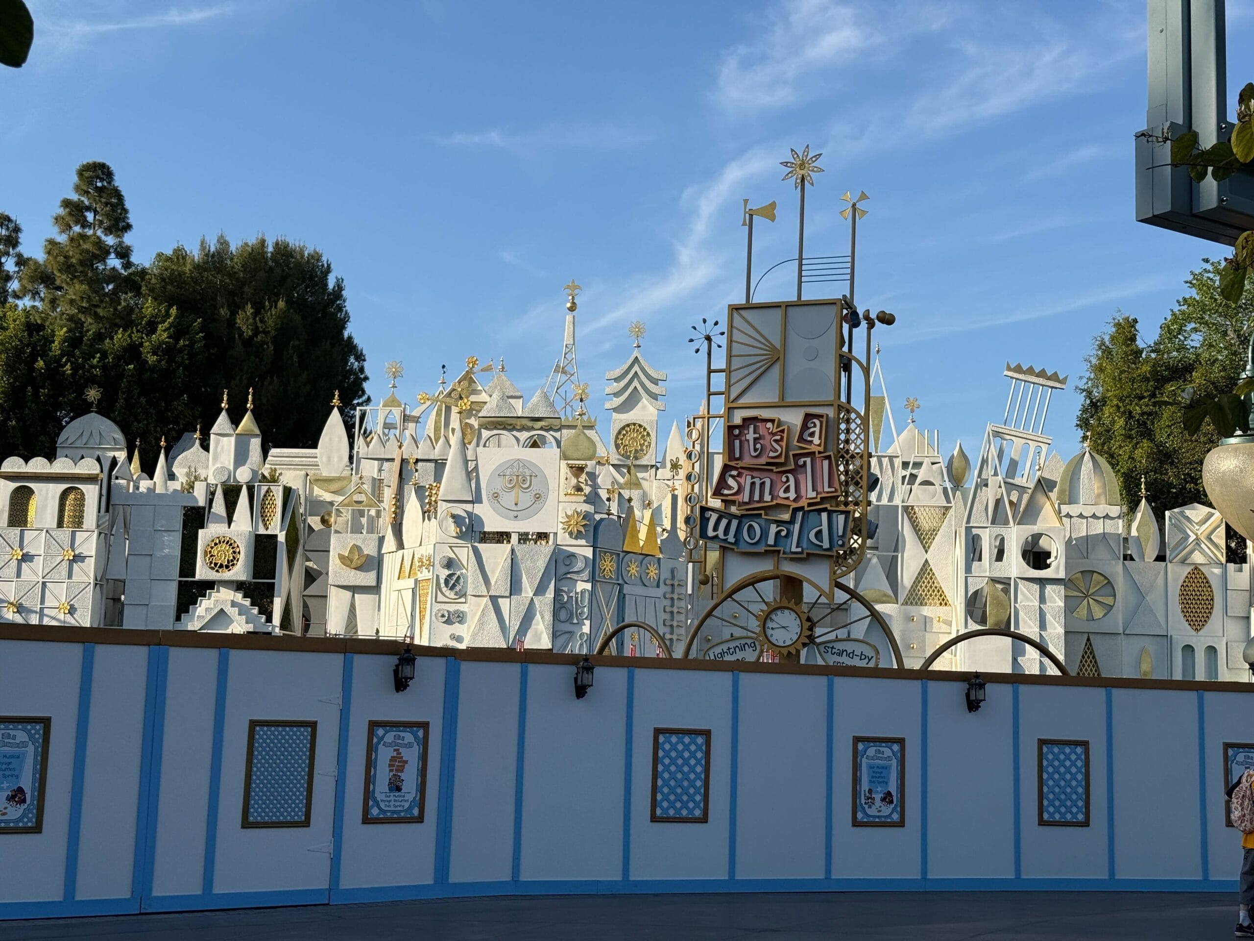 The image captures the exterior of the "It's a Small World" attraction, showcasing its iconic whimsical facade with blue construction walls along the pavement and trees in the background.