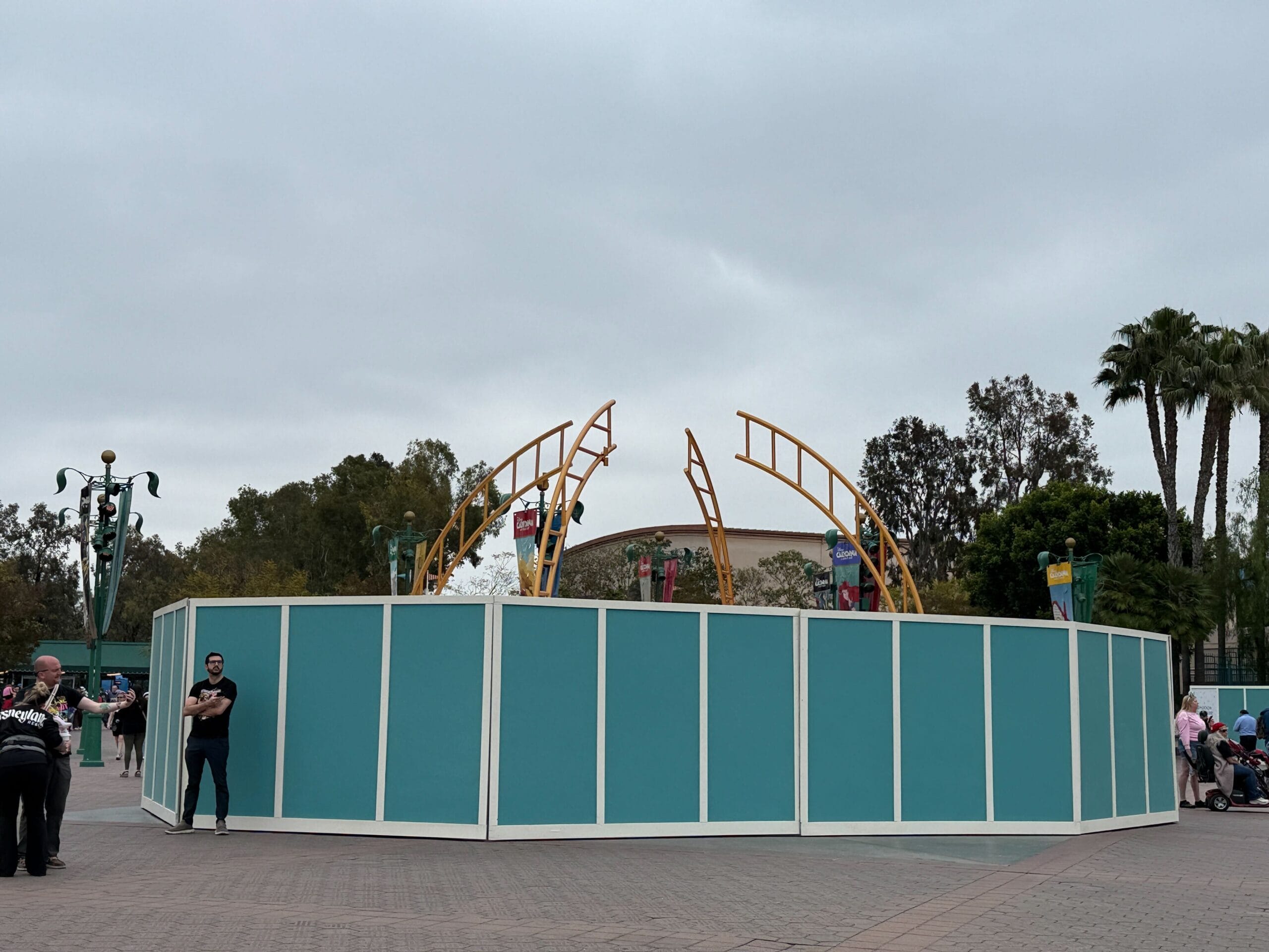 Teal construction walls surround an area with yellow metal arches; people stand nearby under a cloudy sky.