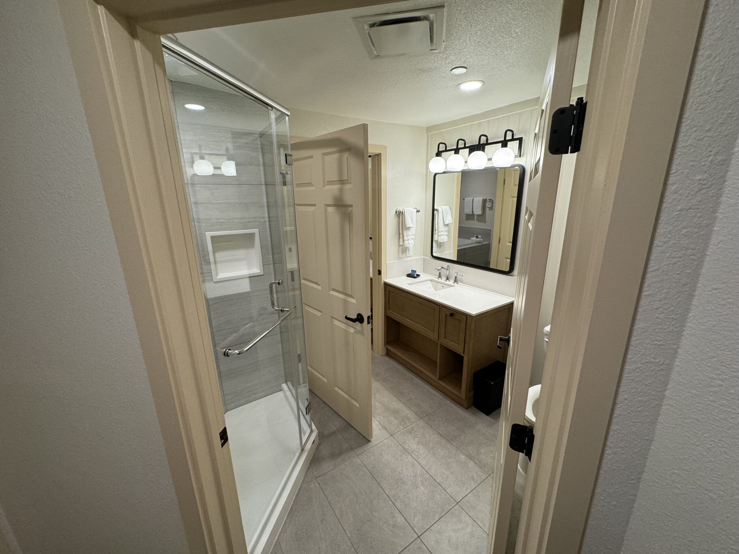 This remodeled villa's modern bathroom features a glass-enclosed shower, white towels, and a large mirror above the vanity. The tiled light gray walls and floor add elegance to this 1-bedroom villa at Disney's Hilton Head Island Resort. The door is partially open, inviting you in.