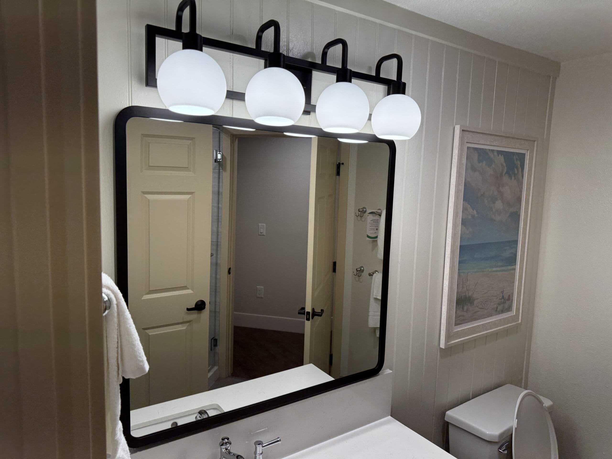 This remodeled villa bathroom features a mirror flanked by four round lights, a cozy towel on the rack, and a charming framed beach picture on the wall. Perfect for unwinding after a day at Disney's Hilton Head Island Resort.