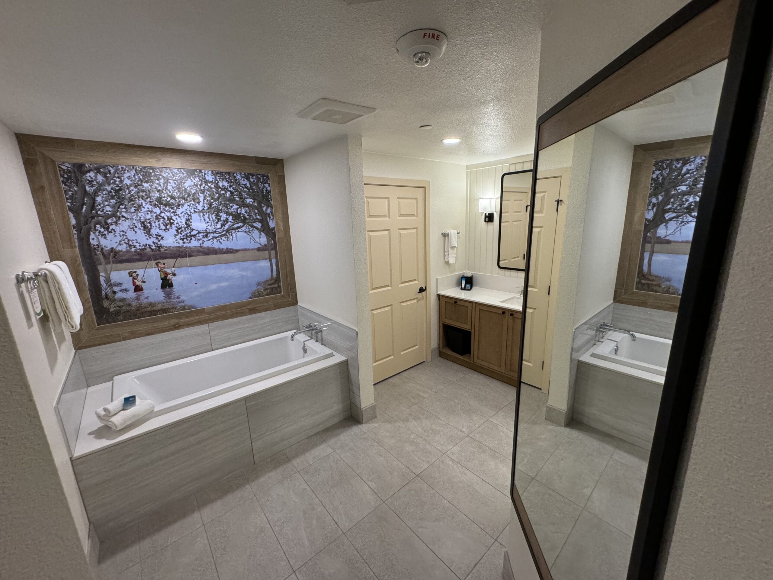 Explore a remodeled 1-bedroom villa featuring a modern bathroom with a large bathtub, mounted towels, framed art above the tub, and a mirror reflecting the serene space. A door leads to another room, inviting you to tour Disney's Hilton Head Island Resort in style.
