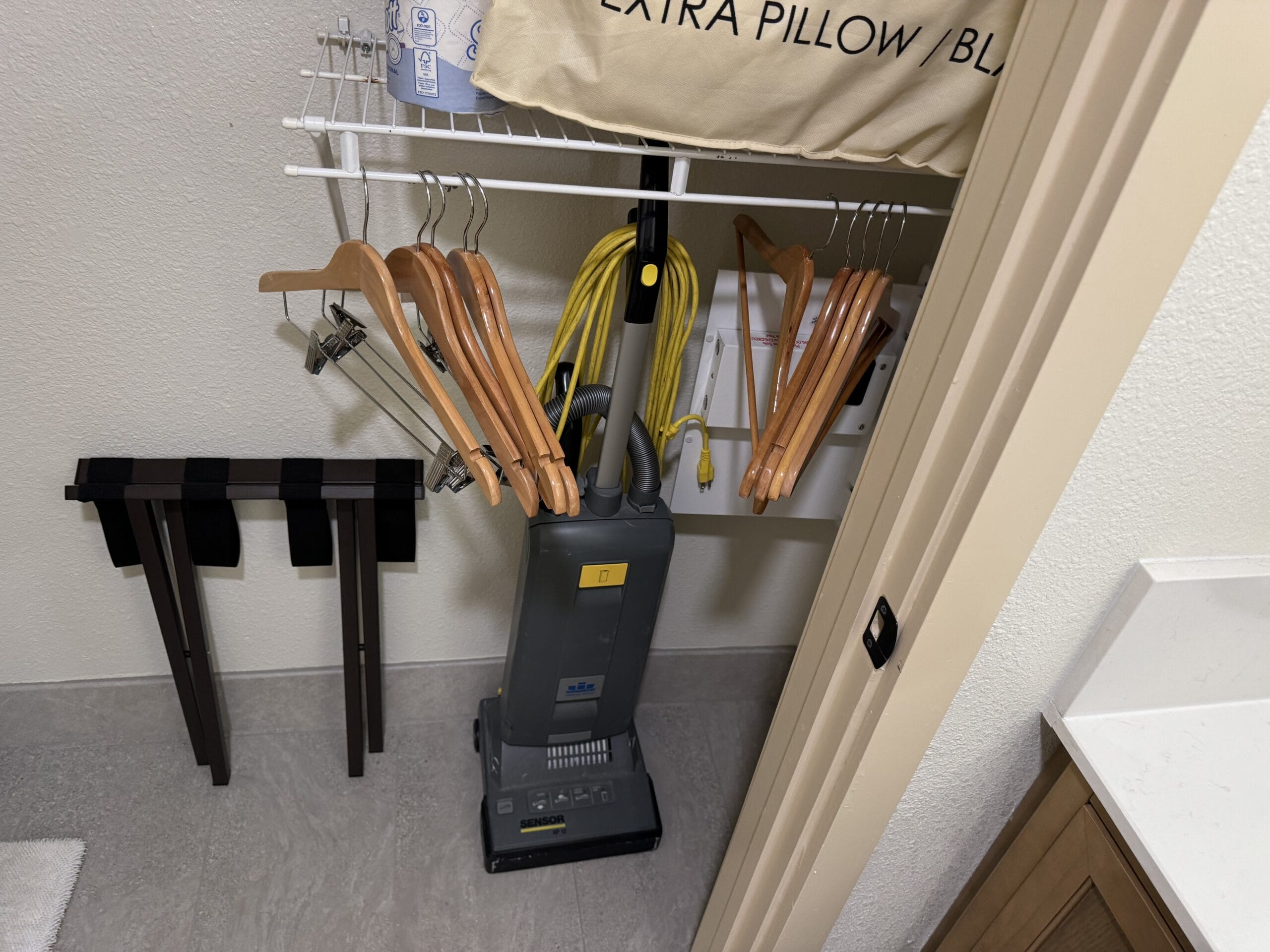 A closet in the remodeled 1-bedroom villa at Disney's Hilton Head Island Resort features hangers, a vacuum cleaner, an ironing board, and a pillowcase with "EXTRA PILLOW" printed on it.