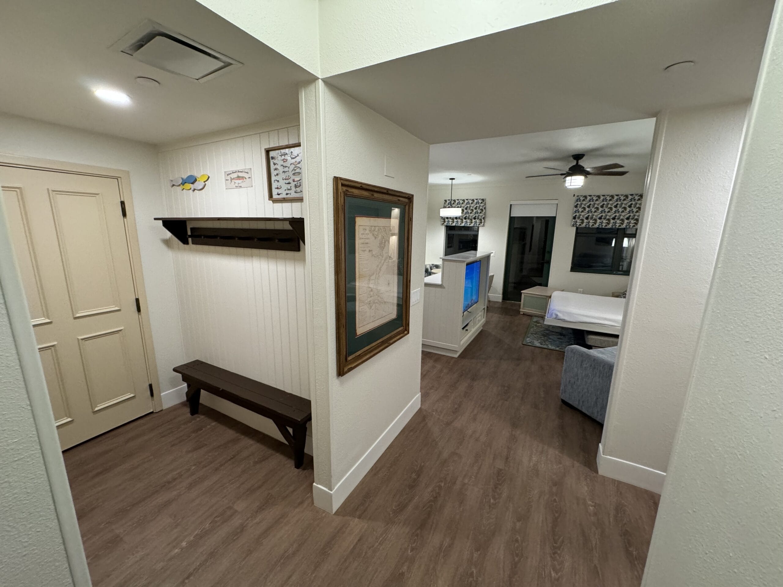 Entryway to a remodeled villa with a wooden bench and coat rack. The room features wood flooring, a ceiling fan, and a TV. A bed is partially visible around the corner in this stylish 1-bedroom villa at Disney's Hilton Head Island.
