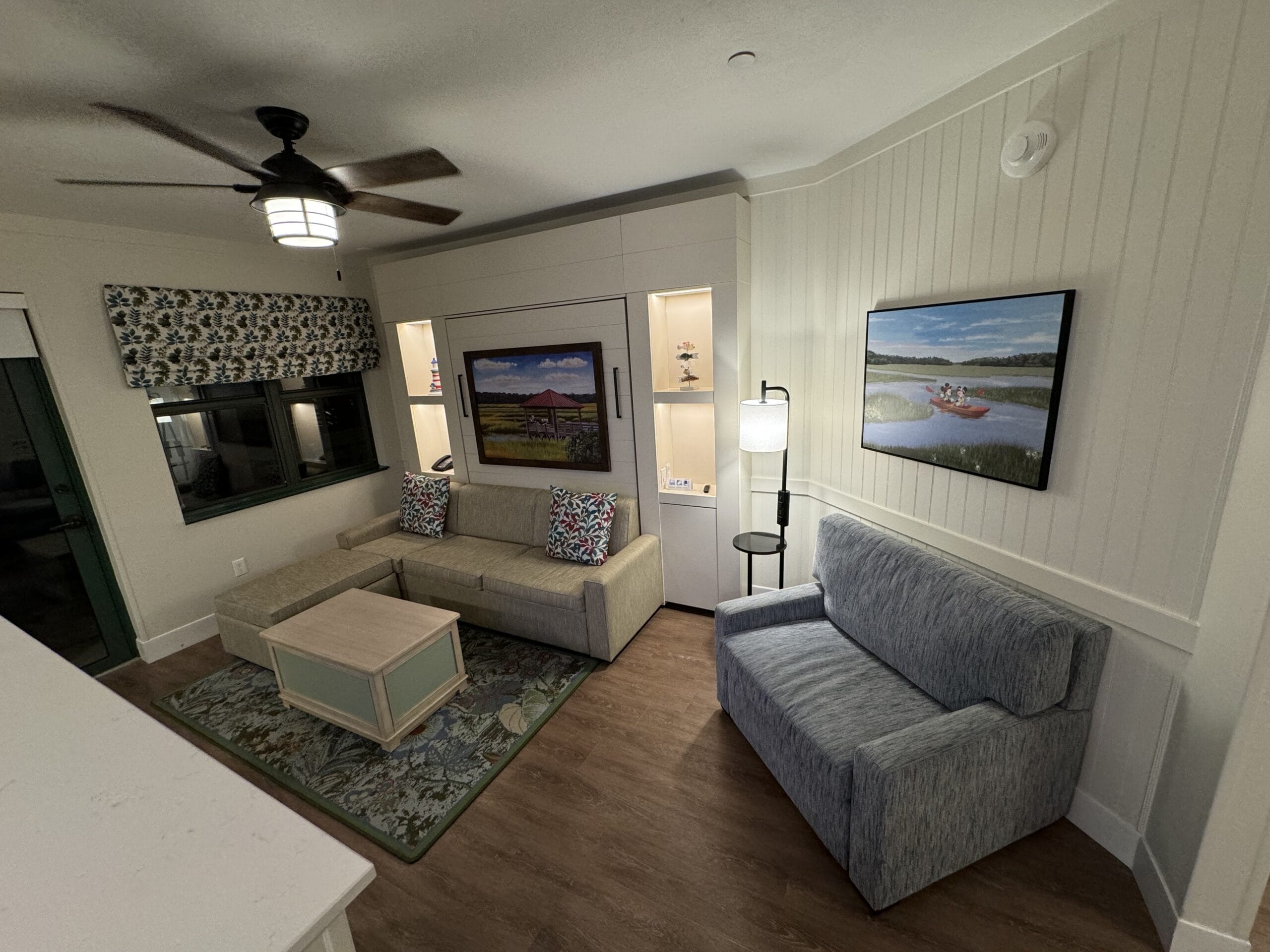 This cozy remodeled 1-bedroom villa features a charming living room with a ceiling fan, sectional sofa, and second sofa. Built-in shelves are adorned with artwork, while a standing lamp adds warmth—perfect for envisioning your next tour at Disney's Hilton Head Island Resort.