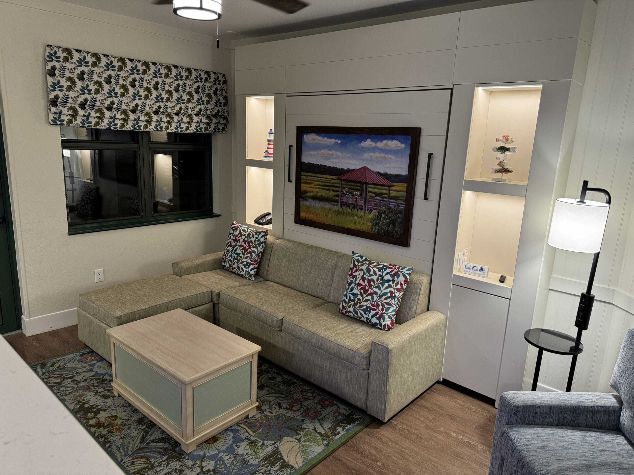 Remodeled 1-Bedroom Villa: Living room with a Murphy bed, gray sofa, floral curtains, decorative throw pillows, built-in shelves, and a floor lamp. A framed painting is above the sofa—a charming retreat at Disney's Hilton Head Island Resort.