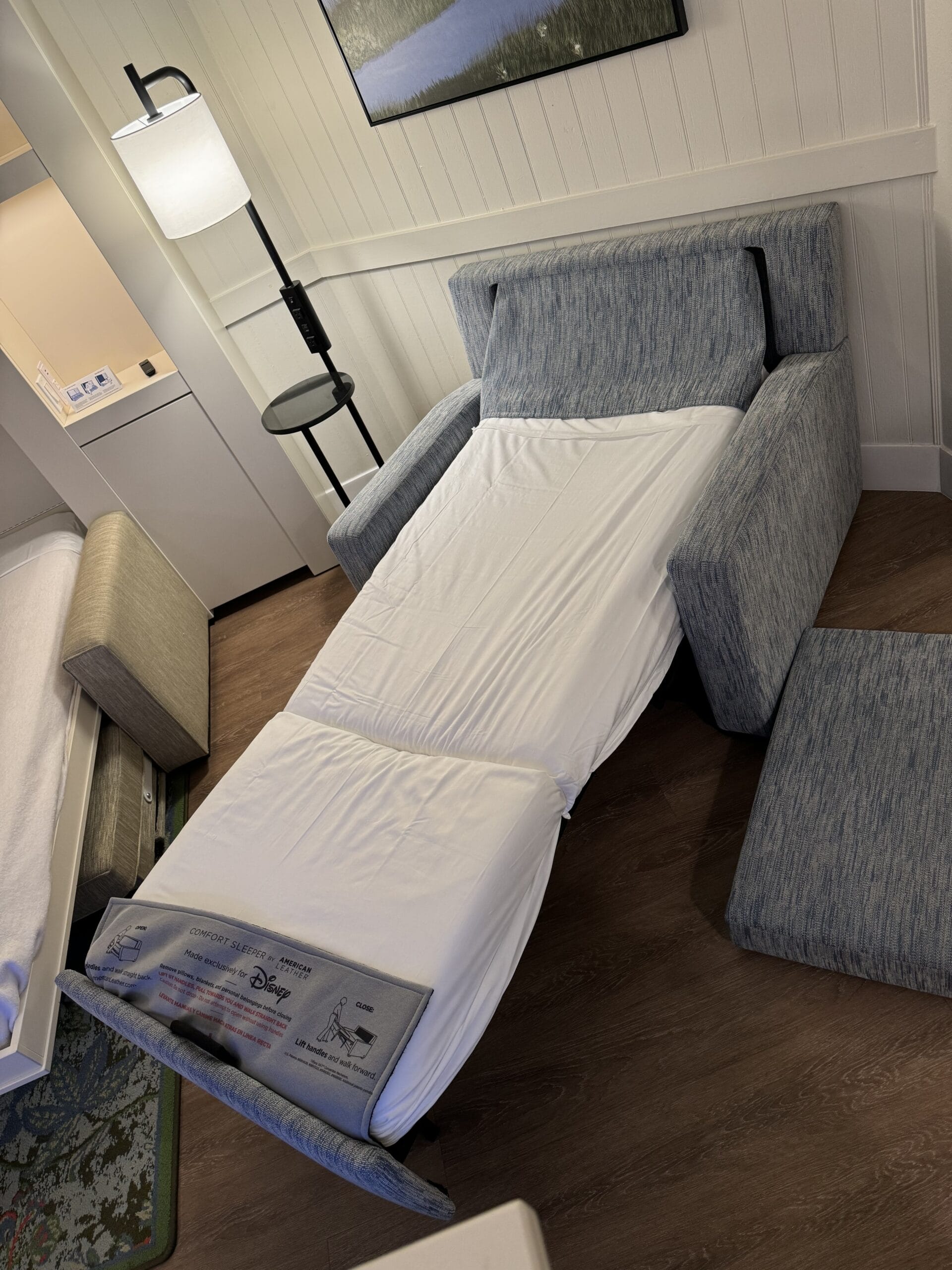 The 1-bedroom villa at Disney's Hilton Head Island Resort features a pull-out sofa bed partially extended in a cozy room with a floor lamp and side table nearby. The bedding is pristine white, complementing the hardwood flooring.