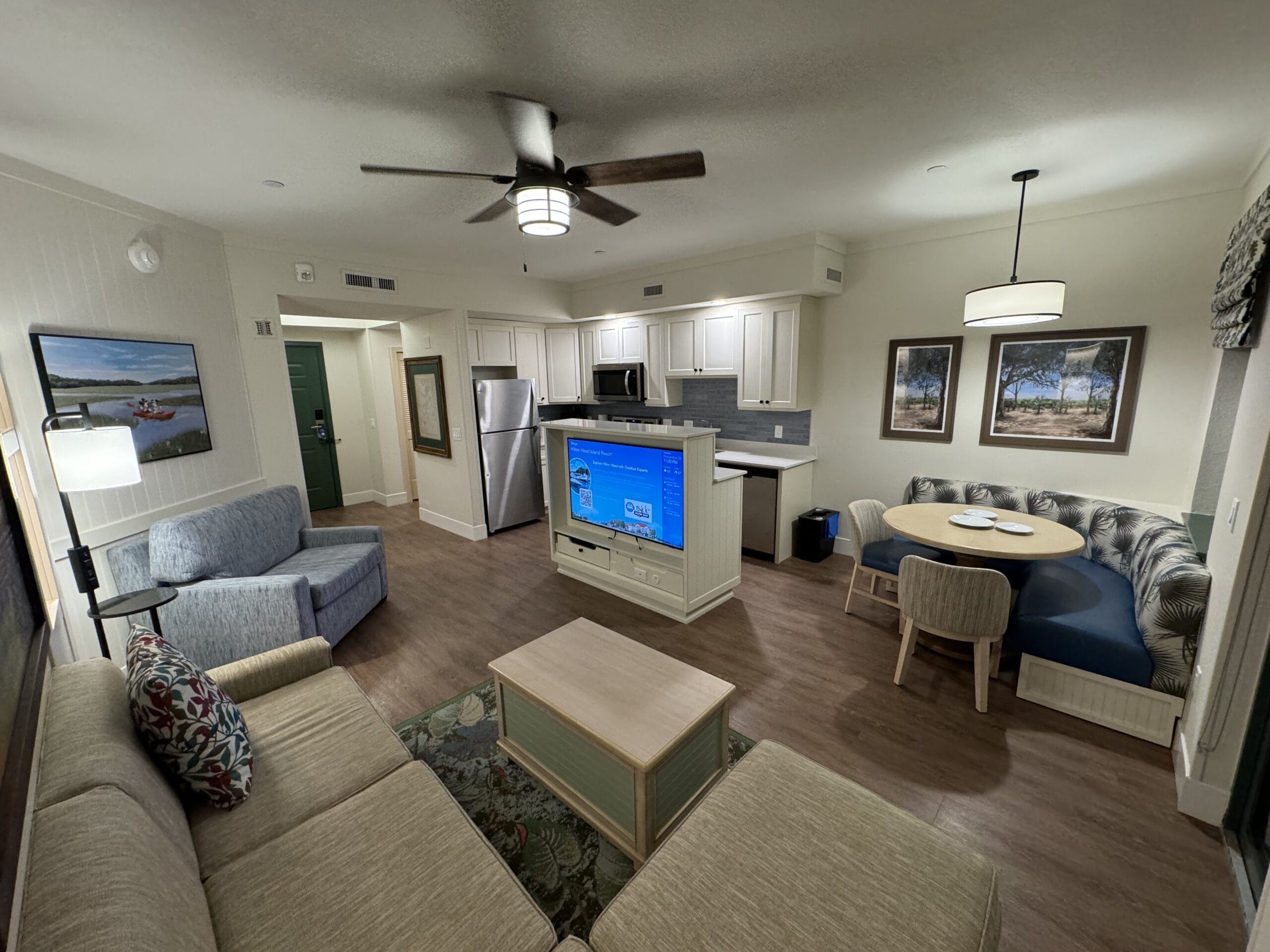 Discover a remodeled 1-bedroom villa featuring a spacious, modern living room with wood floors, ceiling fan, sectional sofa, armchair, and coffee table. Enjoy the nearby dining area with stainless steel appliances at Disney's Hilton Head Island Resort.