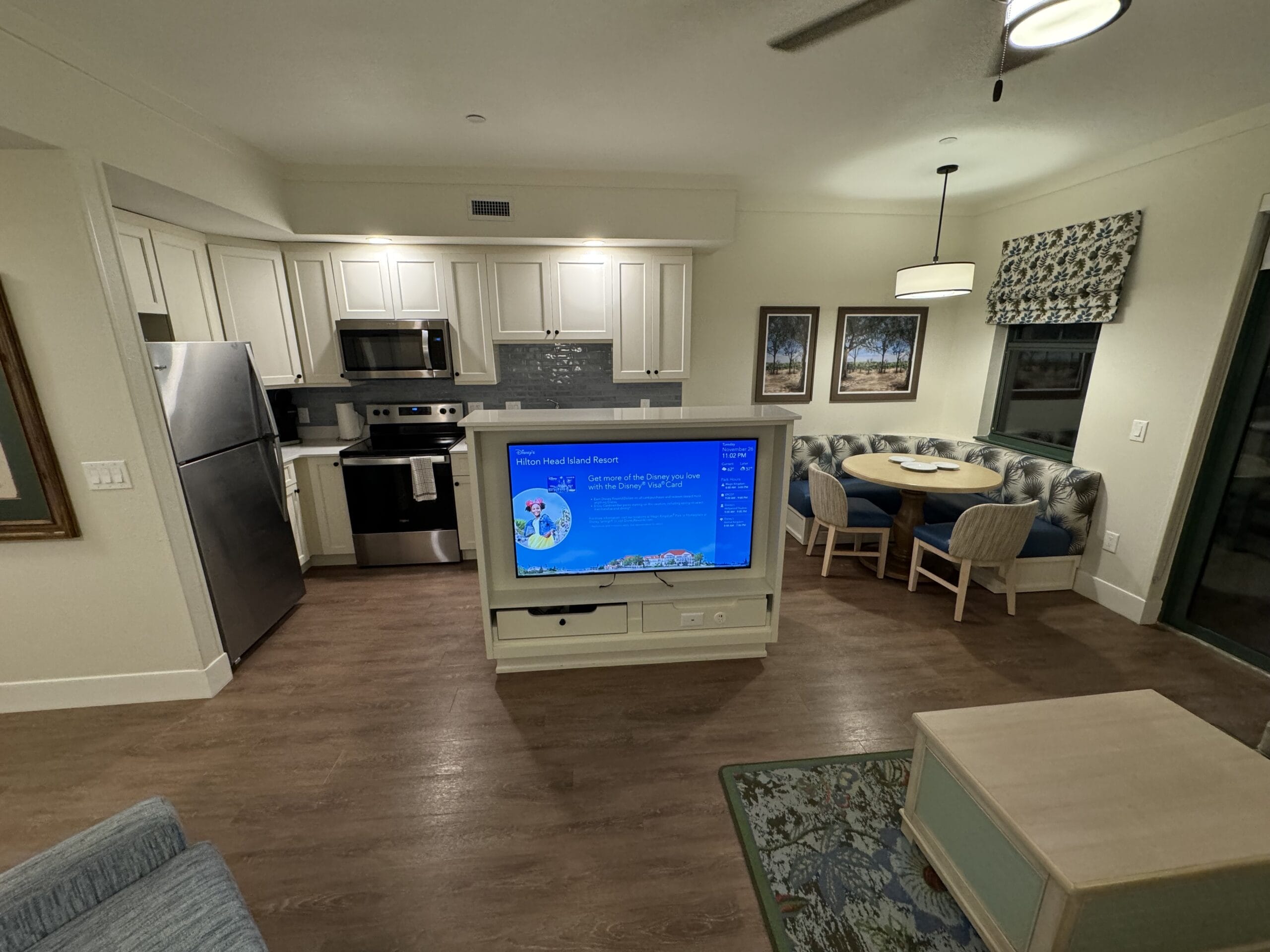 Discover the remodeled 1-bedroom villa featuring a modern open-plan kitchen and living area with a mounted TV, stainless steel fridge, round dining table with chairs, and light wooden flooring—perfect for relaxing after a tour of Disney's Hilton Head Island Resort.