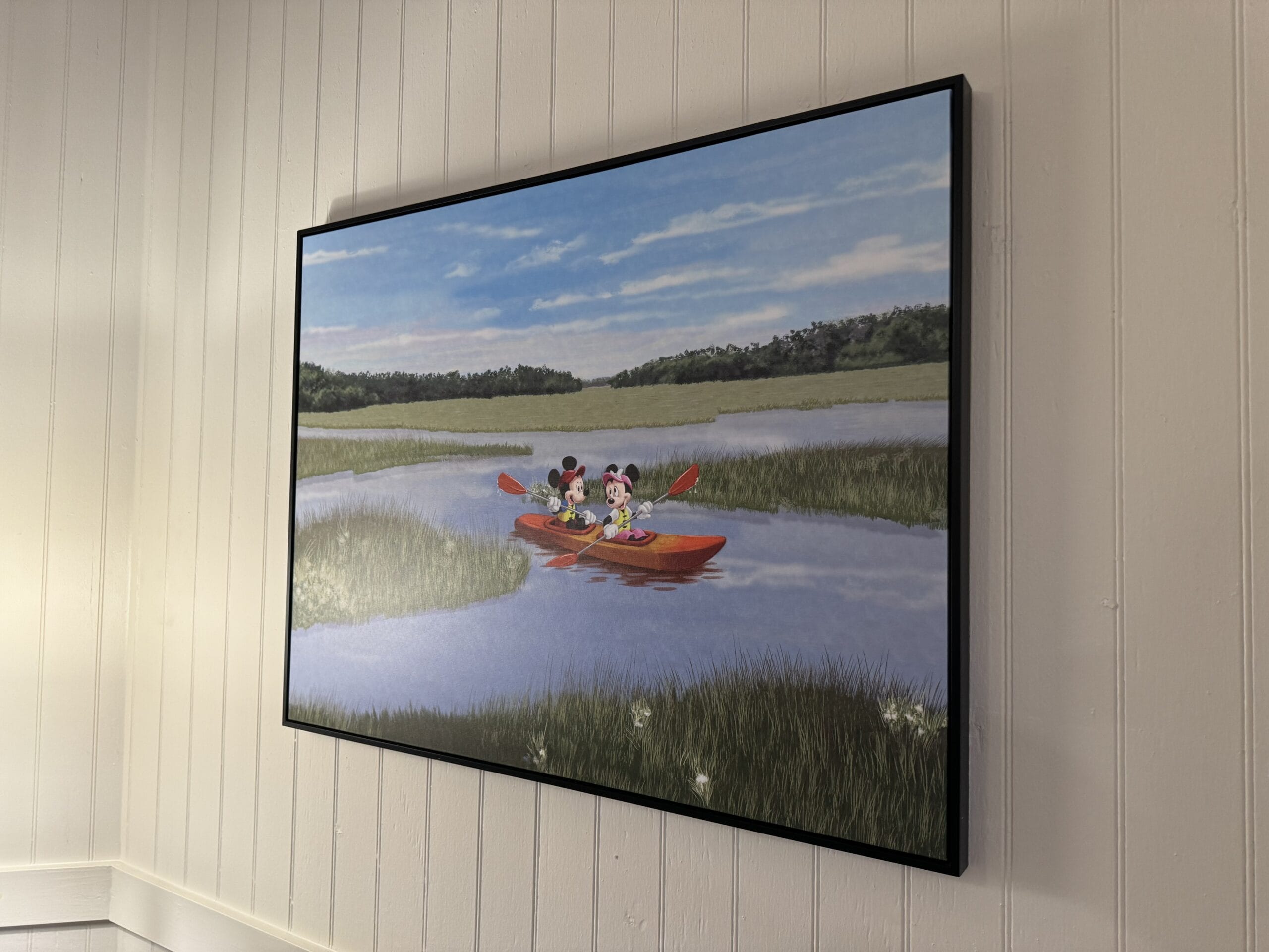 A framed painting on a white wall at Disney's Hilton Head Island Resort depicts two animated characters in a red canoe, paddling serenely on a river with grassy banks under a blue sky, reflecting the whimsical charm of your remodeled 1-bedroom villa.
