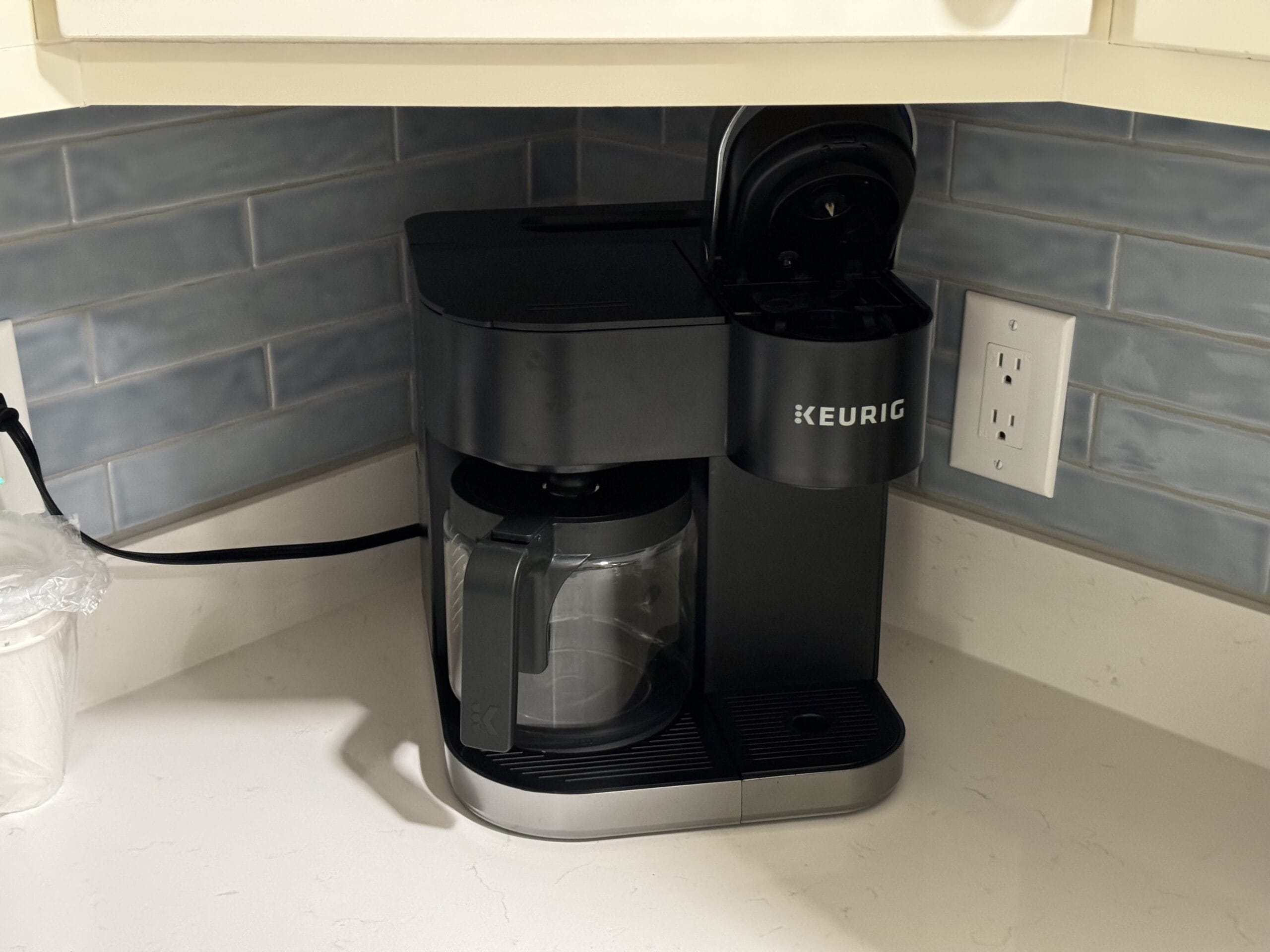 A Keurig coffee maker with an open lid sits on a kitchen counter against a blue tiled backsplash, reminiscent of the charming decor you'd find in a remodeled villa at Disney's Hilton Head Island Resort, conveniently located beside an electrical outlet.