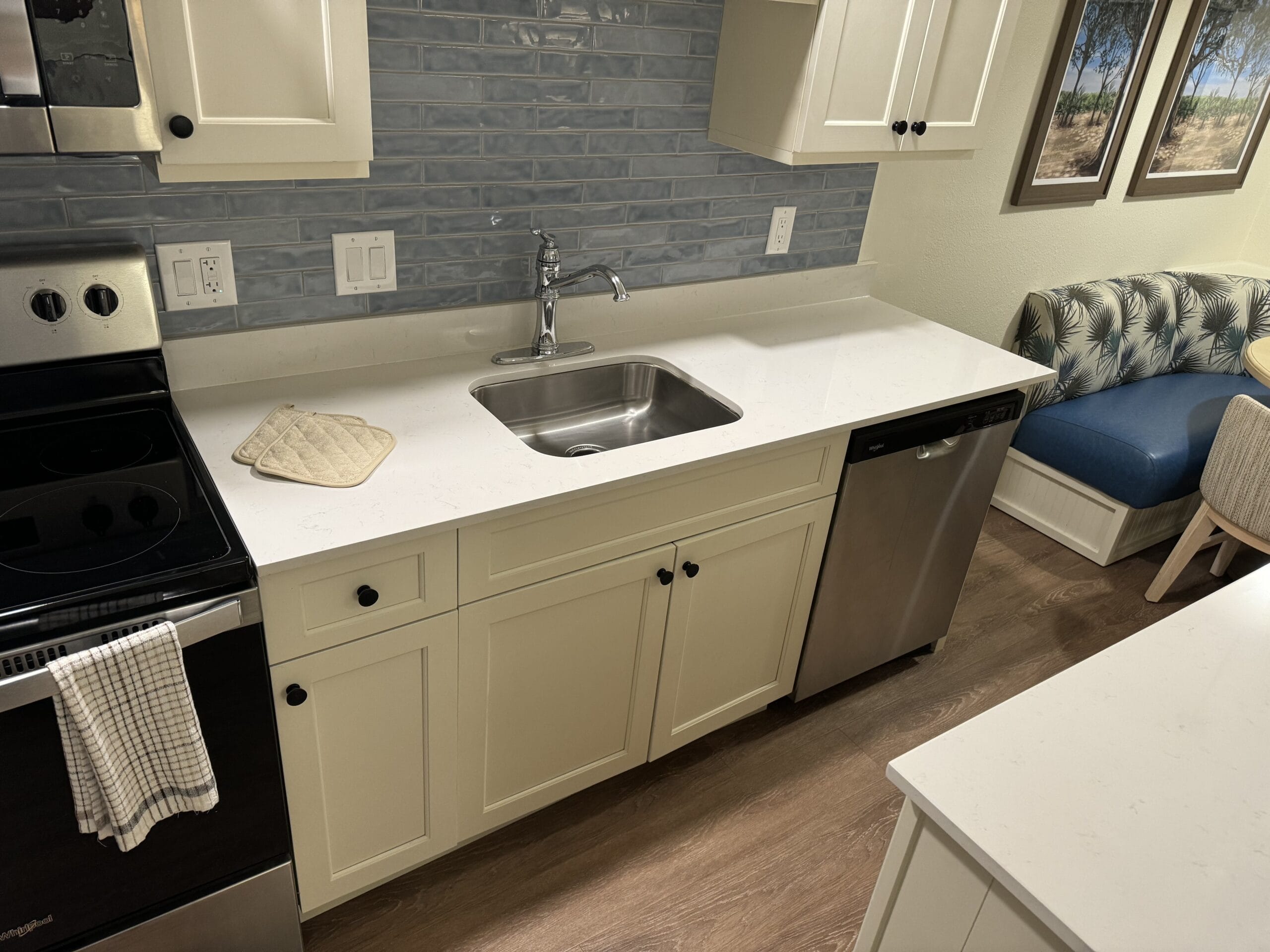 This remodeled 1-bedroom villa features a modern kitchen with white cabinets, stainless steel appliances, and a central sink. A blue tiled backsplash complements the small dining nook with blue cushioned seating. Explore this gem at Disney's Hilton Head Island Resort on a tour.