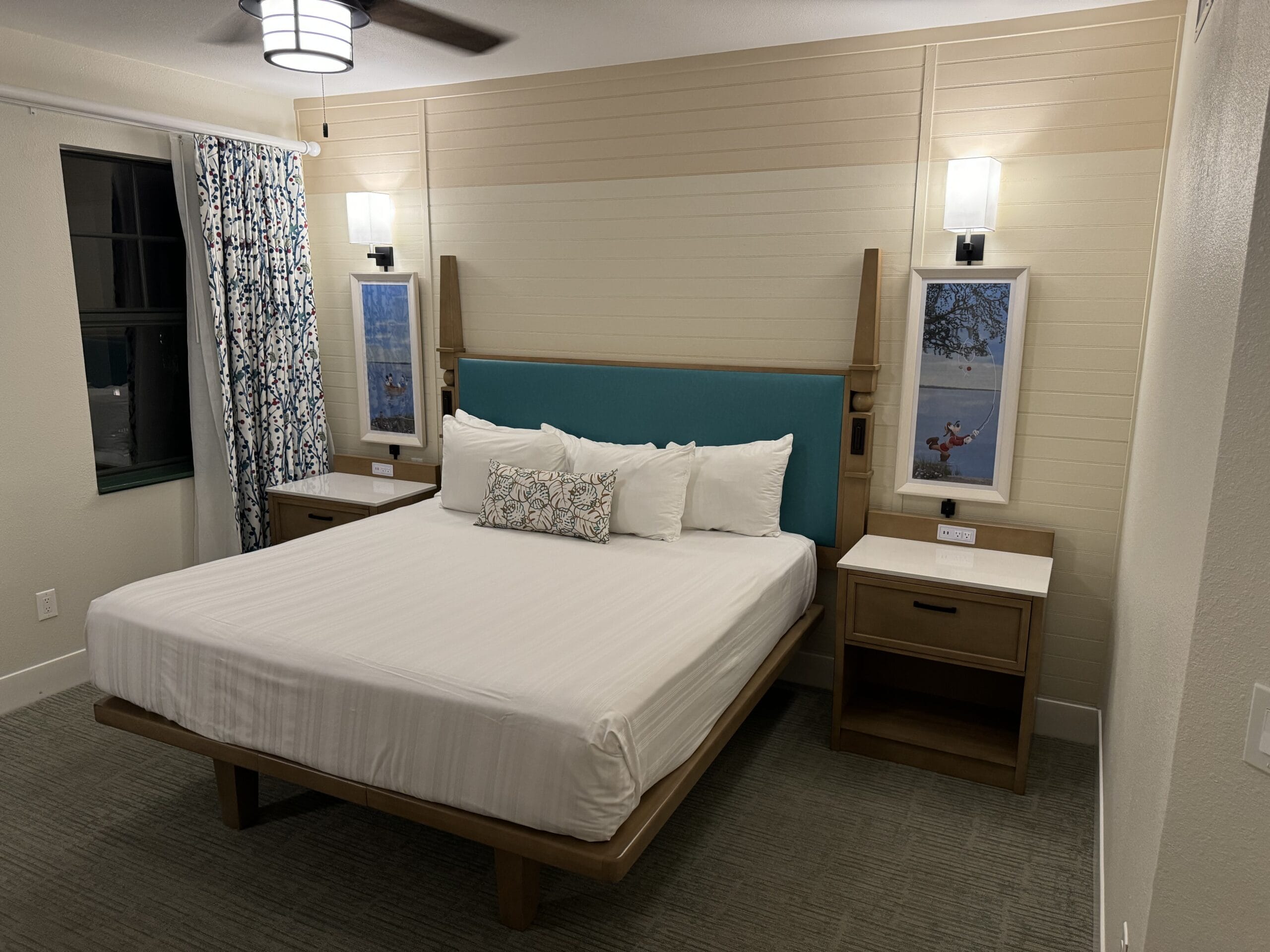 Experience a stay in the remodeled 1-Bedroom Villa at Disney's Hilton Head Island Resort. The room features a large bed with a blue headboard, wooden nightstands, and lamps. Framed pictures adorn the walls, and patterned curtains frame the window, while a cozy rug covers the floor.