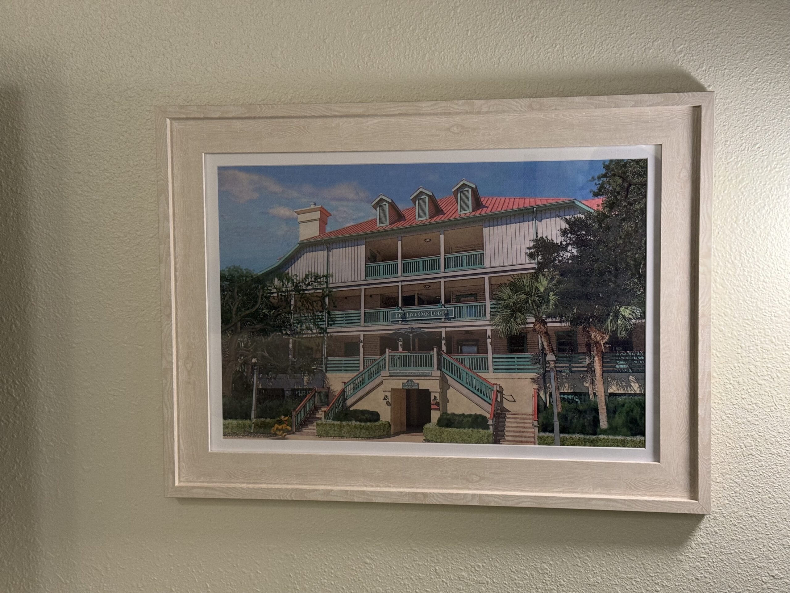 A framed painting of a remodeled villa with several windows, balconies, and a red roof, surrounded by trees and shrubbery, hangs on a light green wall.