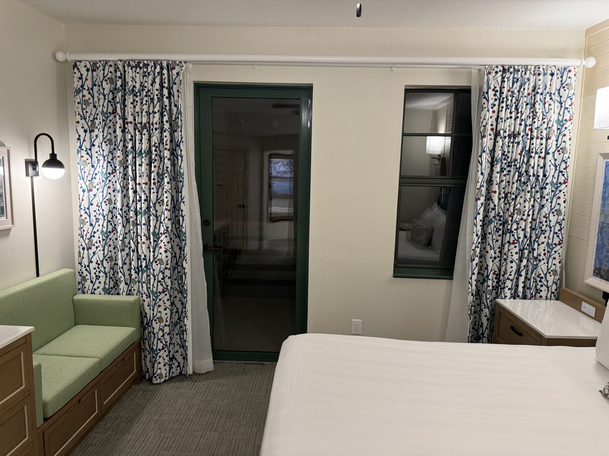 This remodeled 1-bedroom villa at Disney's Hilton Head Island Resort features a cozy bed, green cushioned seating area, patterned curtains, and a glass door. A lit wall lamp casts a warm glow, while the window reflects part of the enchanting room. Ideal for a tour-worthy stay.