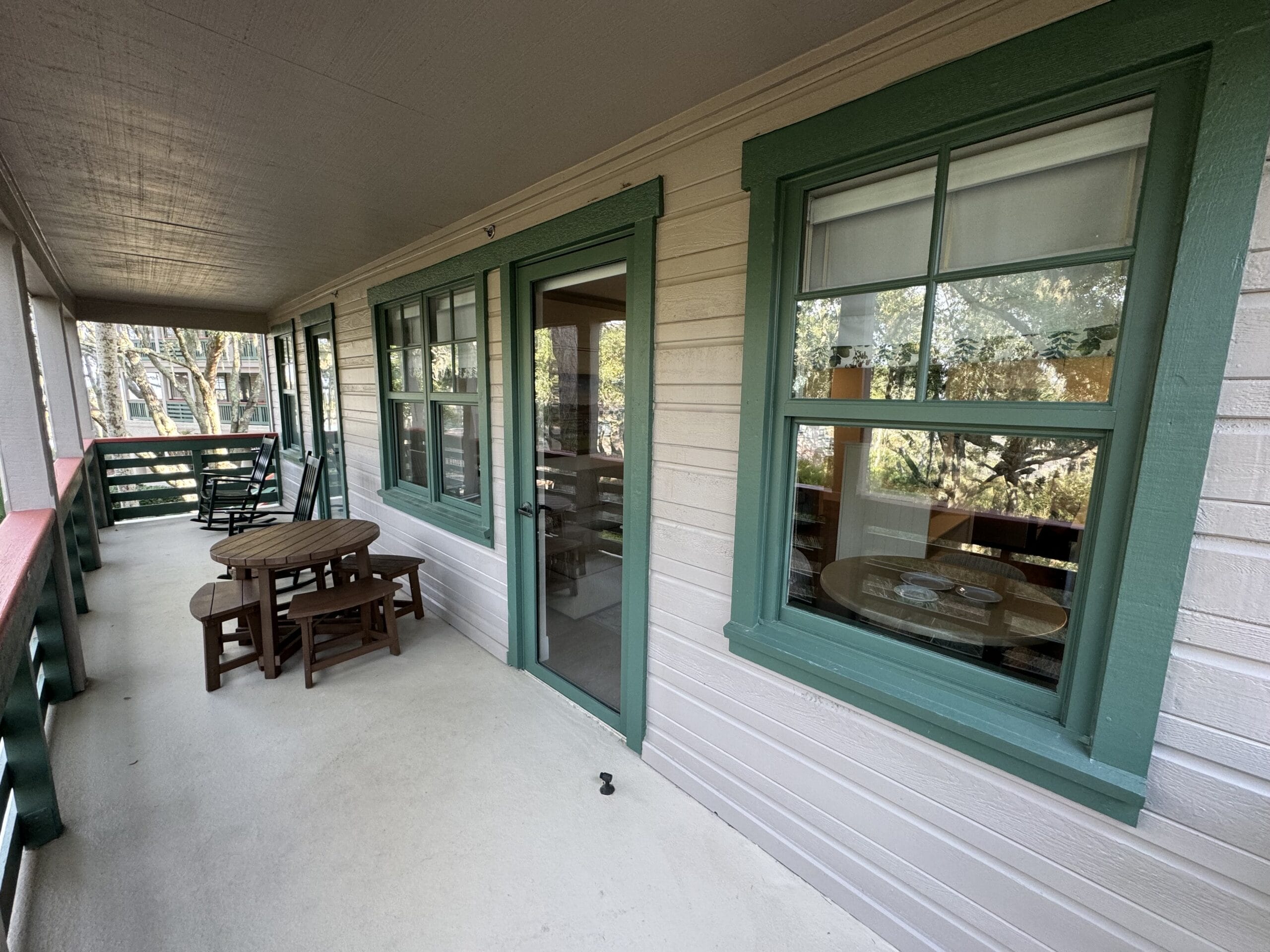 Explore a charming, remodeled 1-bedroom villa featuring a covered porch with green-framed windows, a glass door, and outdoor furniture including a round table, chairs, and a rocking chair. Experience the serene setting reminiscent of Disney's Hilton Head Island Resort on your tour.