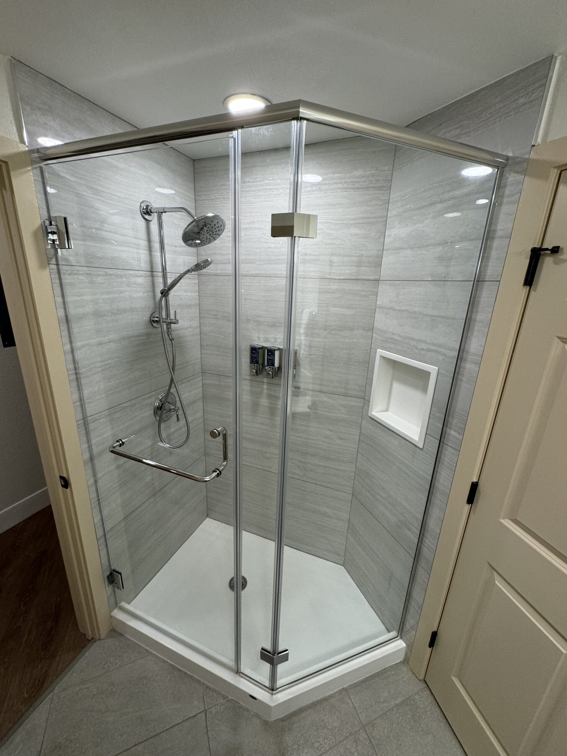 The remodeled villa boasts a modern, glass-enclosed corner shower with a gray tile interior. It features a handheld showerhead, built-in niche, and metal grab bar. The open doors reveal light gray floor tiles, reminiscent of the elegant style at Disney's Hilton Head Island Resort.