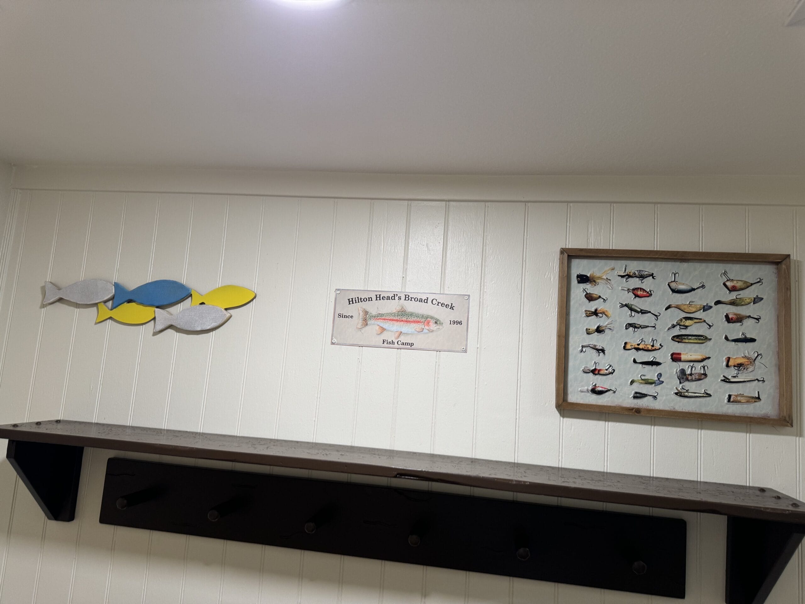 Wall decor in this remodeled villa features a fish theme: three fish cutouts, a framed sign celebrating fishing on Hilton Head and Broad Creek, and another frame showcasing various fish illustrations, all displayed above a wooden shelf with hooks. Perfect for your 1-bedroom villa at Disney's Hilton Head Island Resort.